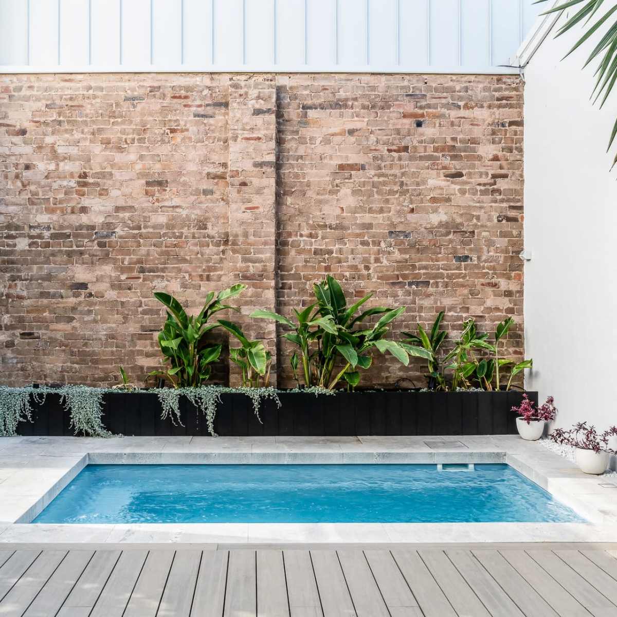 A small swimming pool set against a brick wall, surrounded by potted plants and a wooden deck.