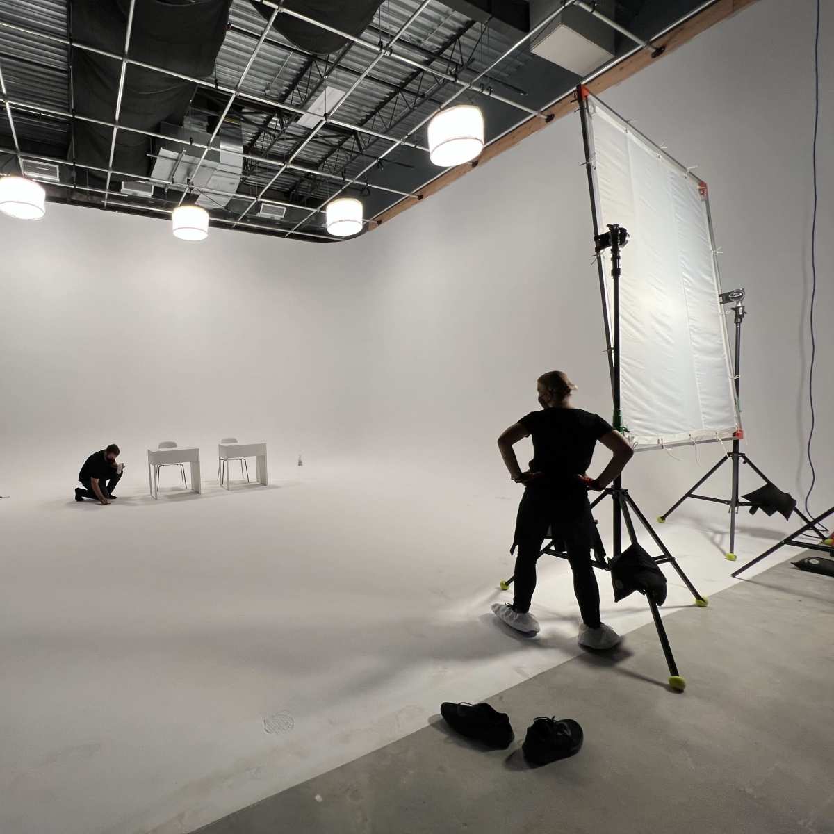 A large, empty studio space is being prepared for a shoot, with two tables and several crew members working in the scene.