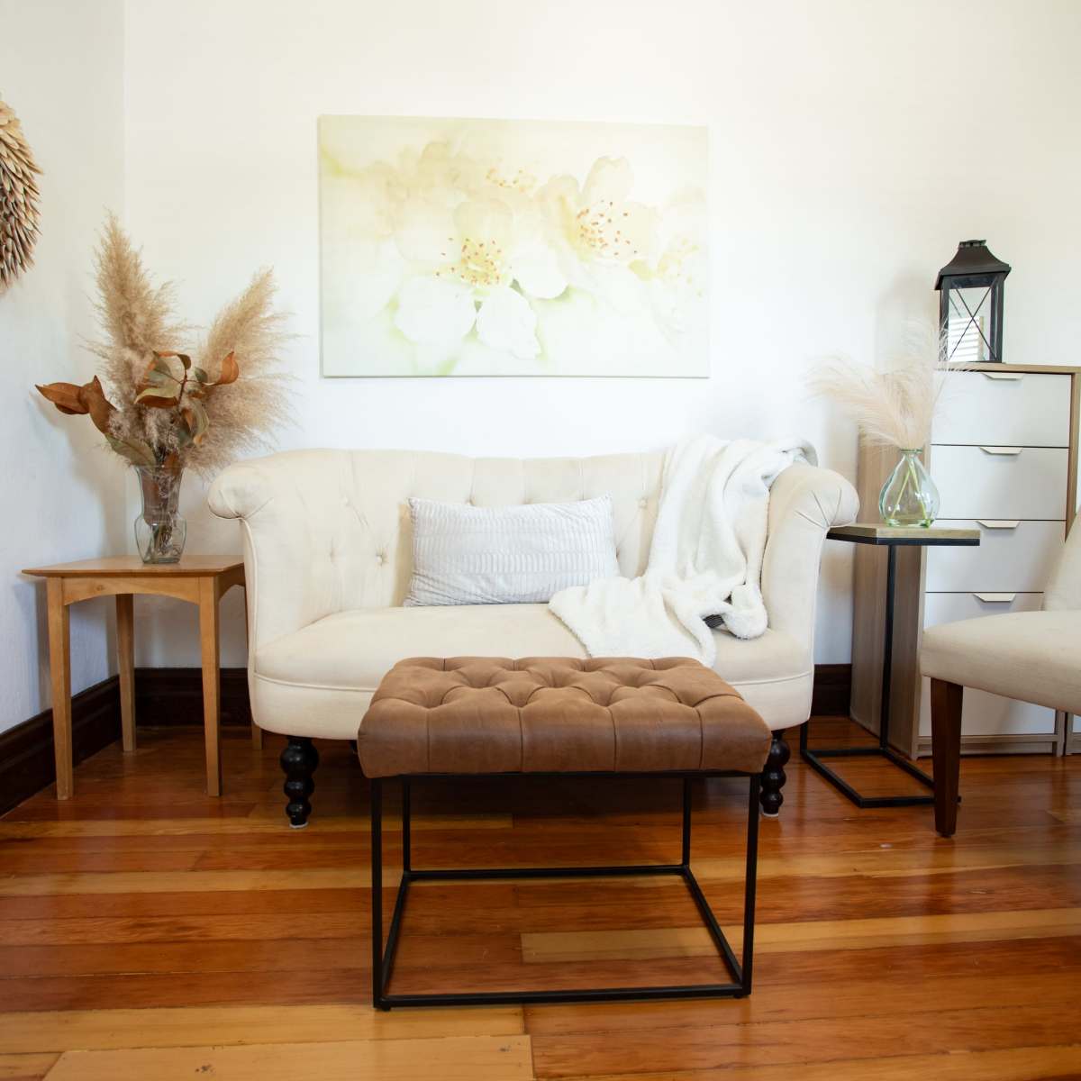 A light and airy living room features a beige tufted ottoman, a white sofa with a throw blanket, and decor elements such as a vase with dried flowers and a small side table.