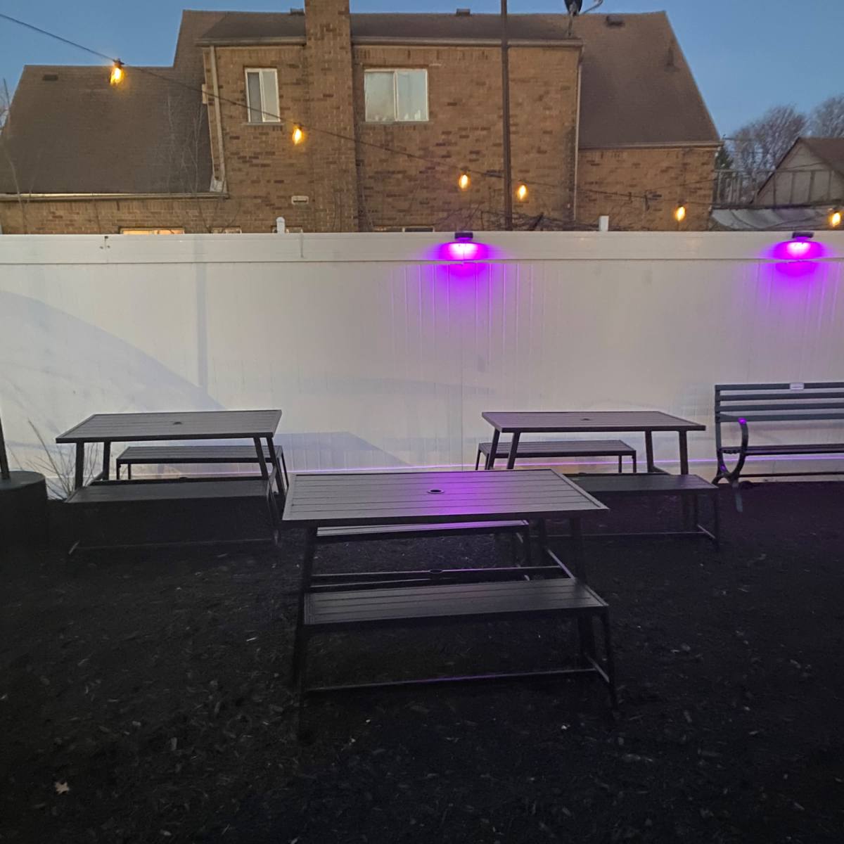Empty metal tables arranged in an outdoor area near a white fence illuminated by purple lights.