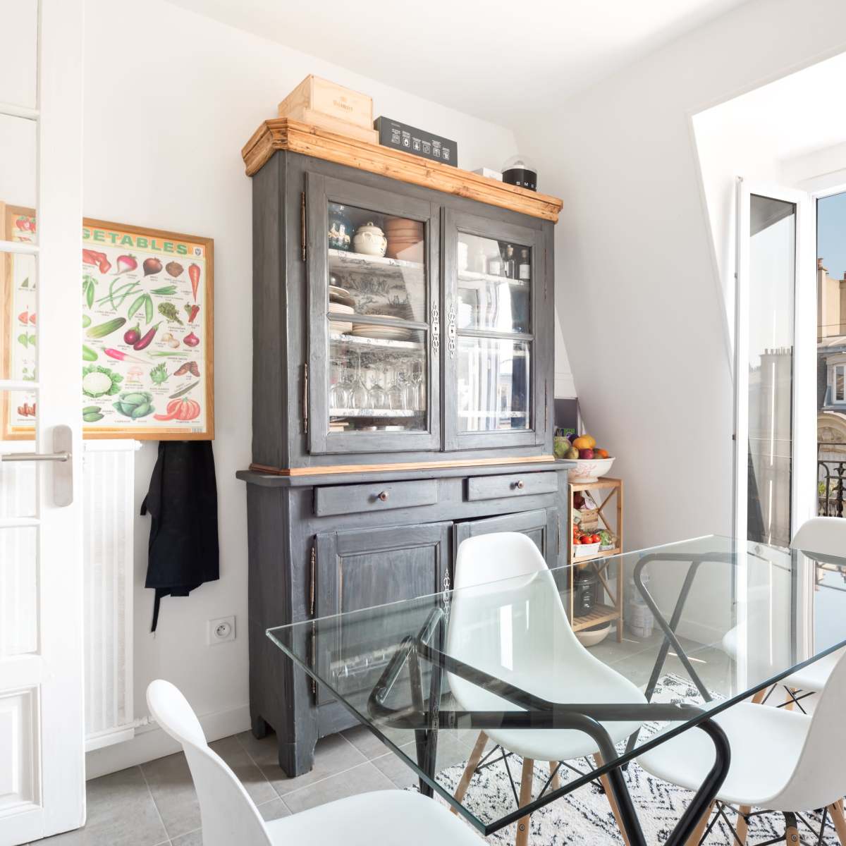 A dining area features a glass table surrounded by white chairs, with a wooden cabinet displaying dishes and a bright window overlooking a balcony.
