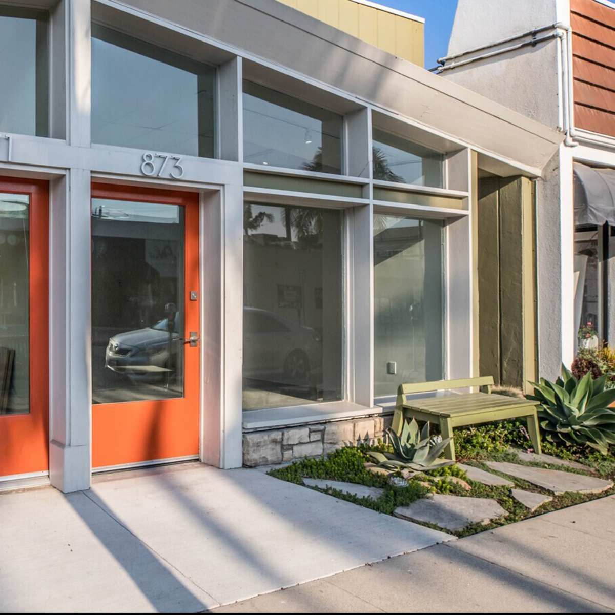 A modern commercial building with large glass windows and orange doors, surrounded by a neatly landscaped area with a bench and stone pathway.