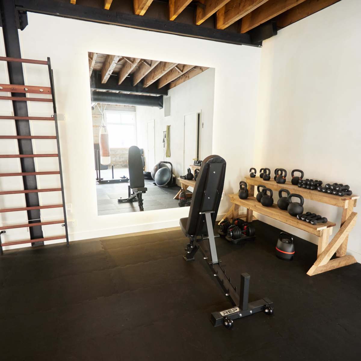 A home gym features a weight bench, a wall-mounted ladder, a mirror, and a rack of kettlebells against a white wall.