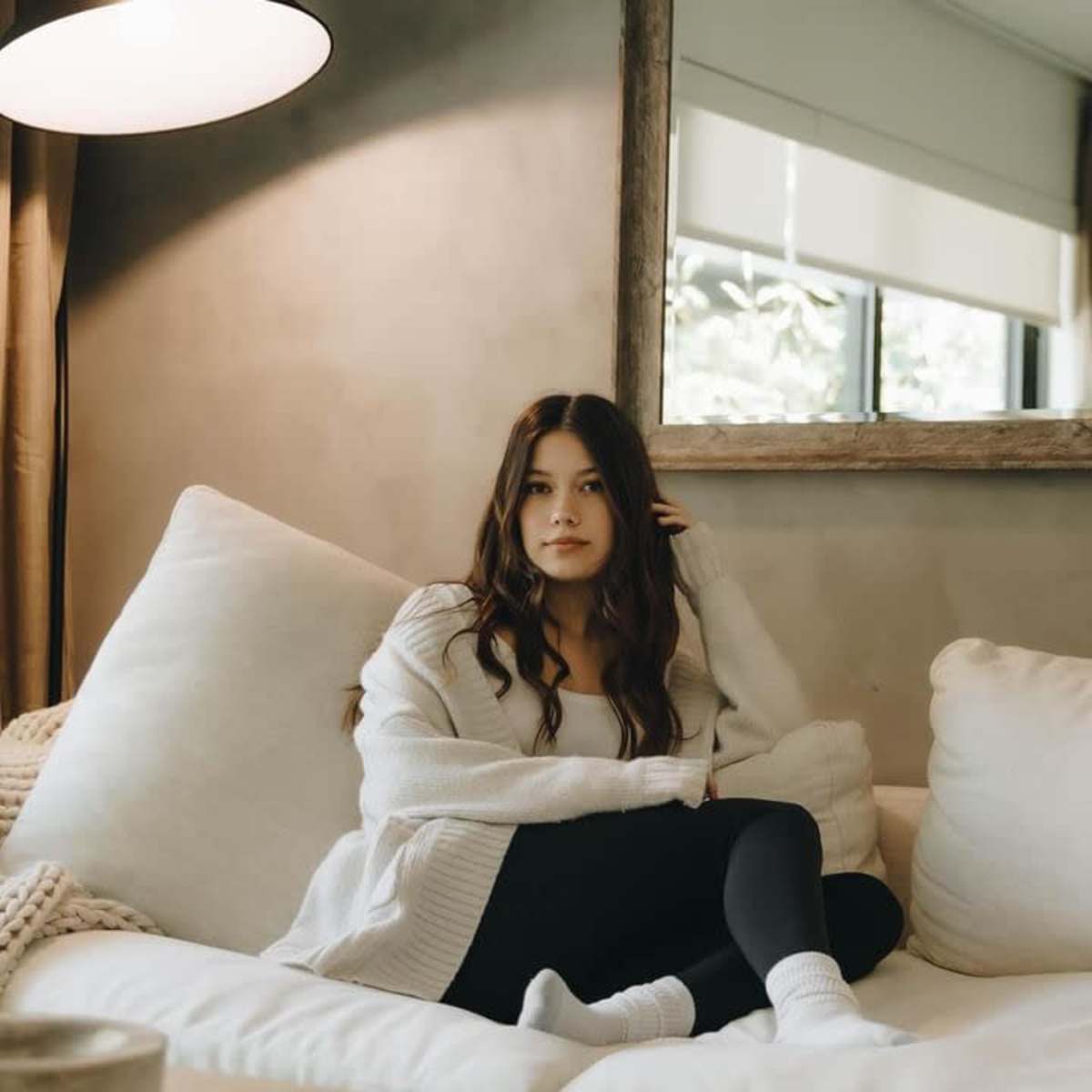 A woman sits on a large white sofa in a cozy living room, with a lamp and a mirror in the background.