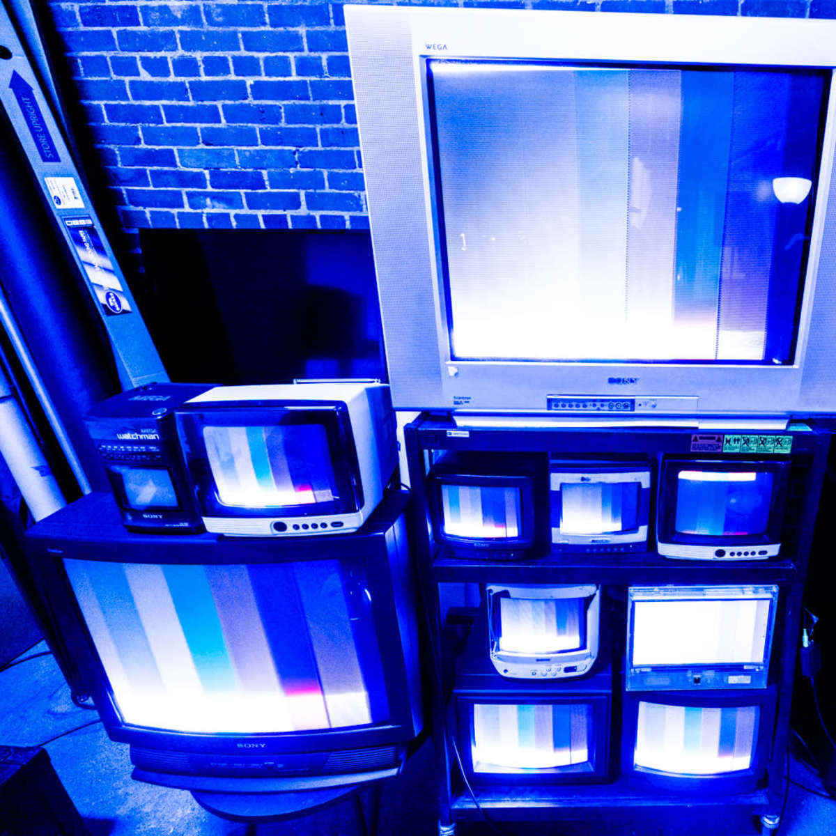 A collection of vintage televisions displaying static and bright vertical lines is arranged against a brick wall.