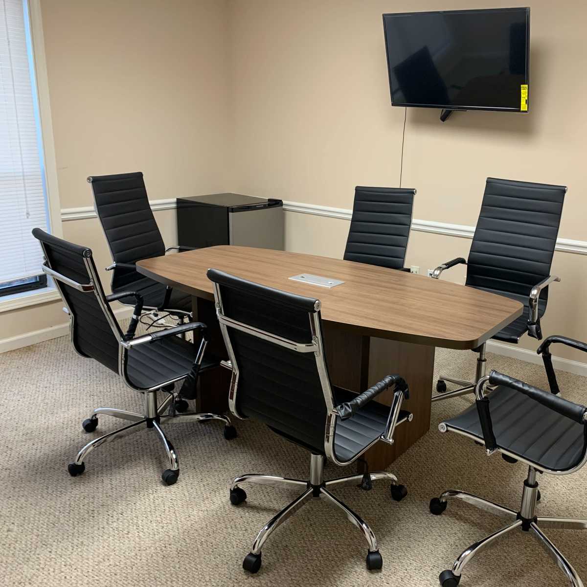 A conference room is furnished with a rectangular wooden table surrounded by six black rolling chairs and features a wall-mounted television.