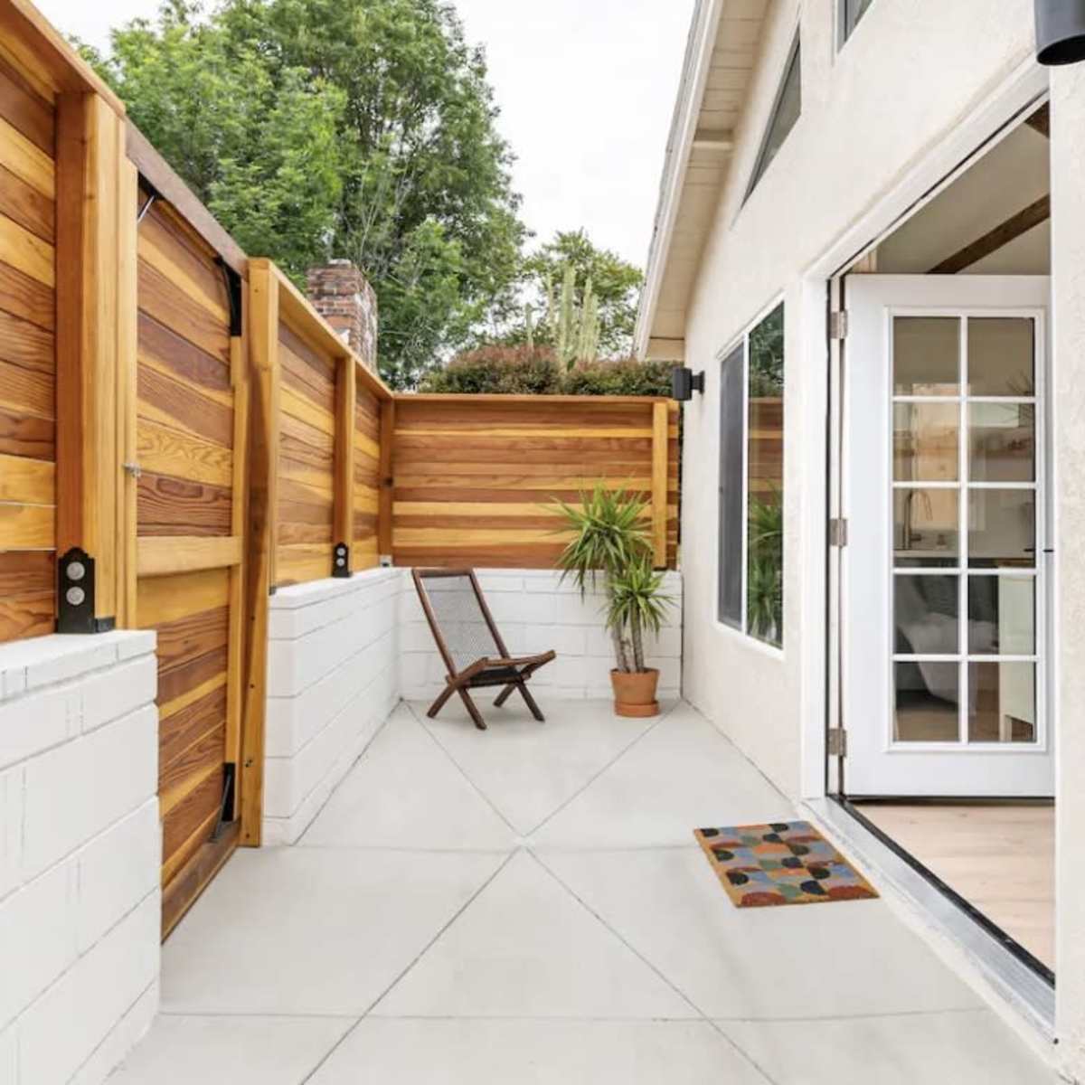 A small patio area enclosed by wooden fences, featuring a potted plant and a folding chair next to a door with French-style windows.