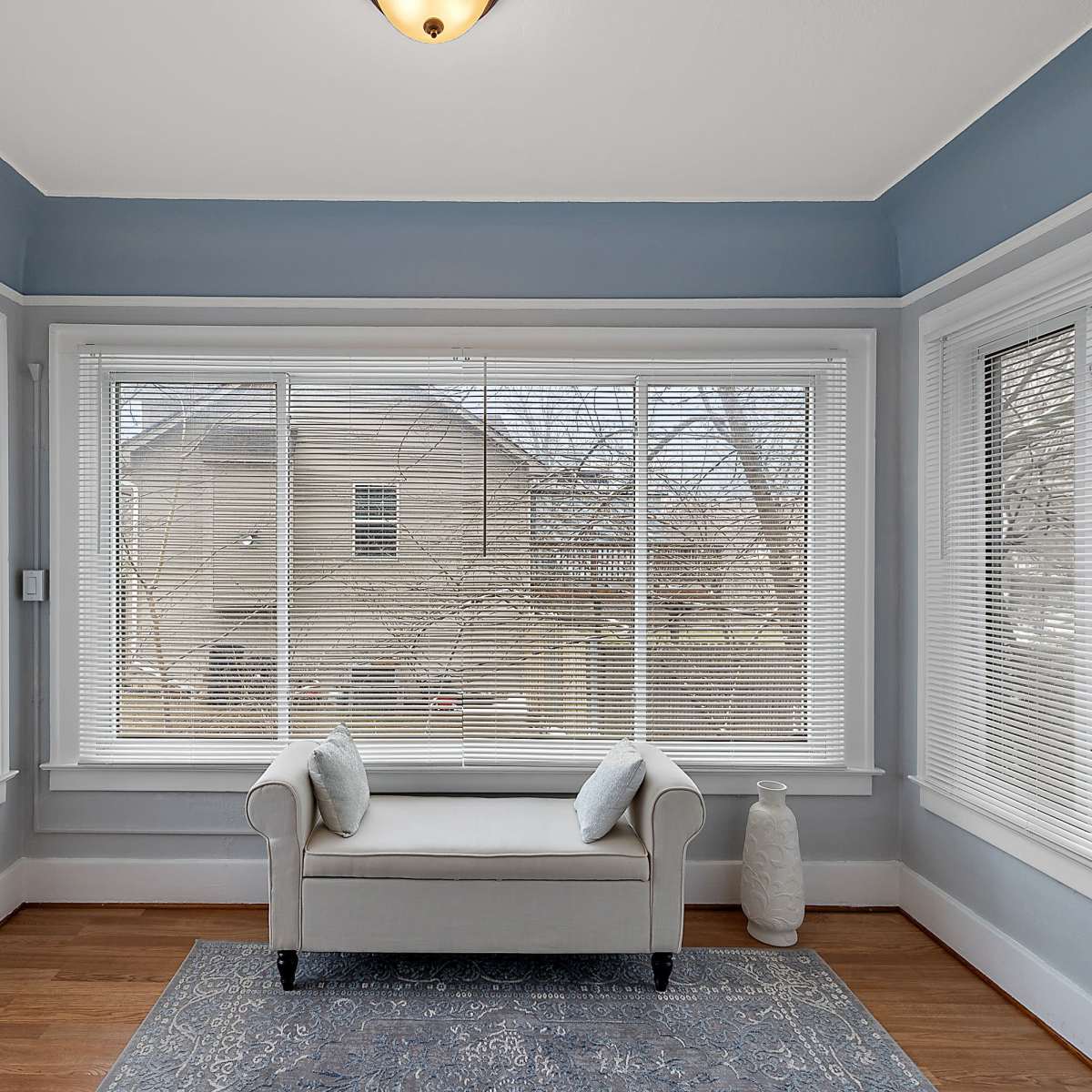 A sunroom features a light-colored bench with two cushions, positioned under a ceiling lamp and large windows covered with horizontal blinds.