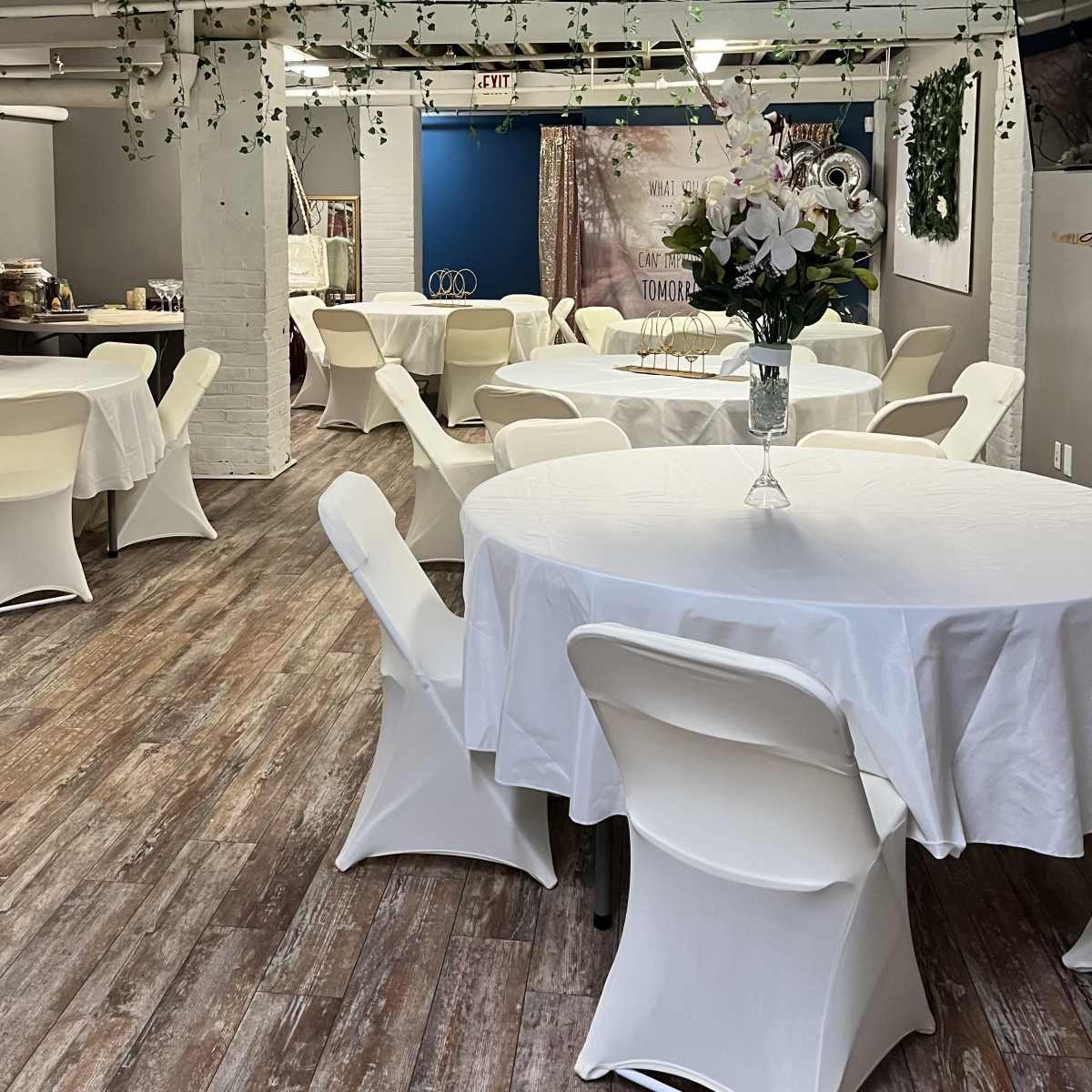 A spacious event room features white tables covered with tablecloths and surrounded by chairs, with decorative plants hanging from the ceiling.