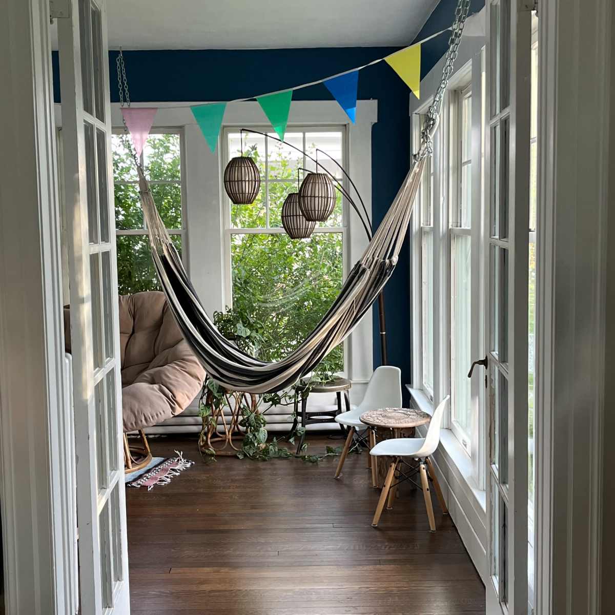 A hammock is suspended between two walls in a bright room filled with plants and decorated with colorful bunting and hanging light fixtures.