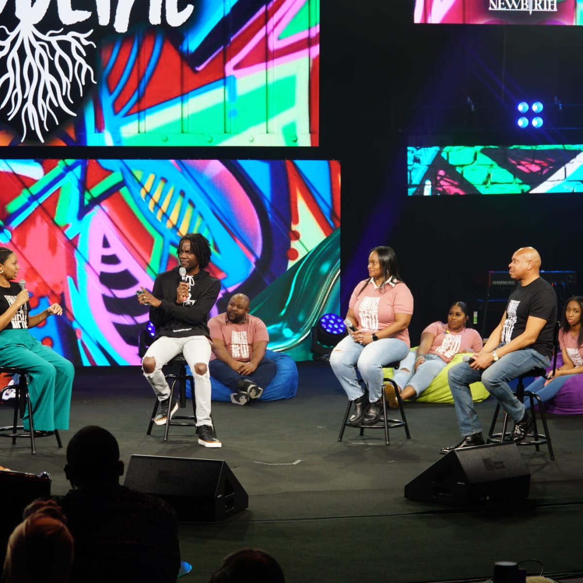 A panel discussion takes place on stage with speakers seated on stools in front of a colorful backdrop, while several participants sit on the floor behind them.