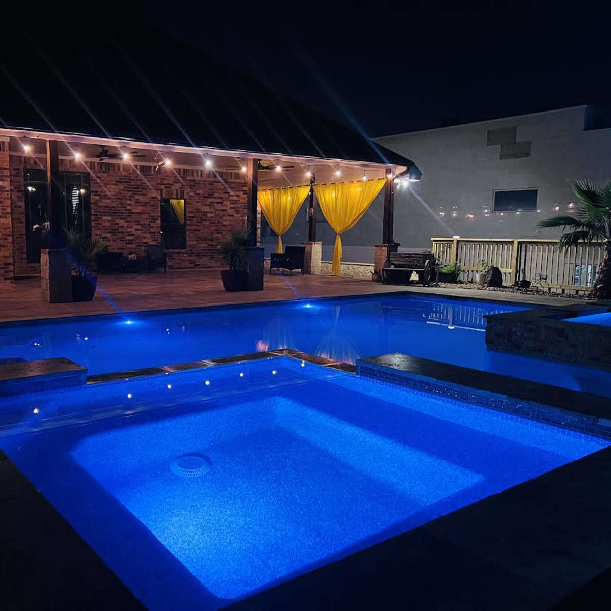 The scene features a well-lit backyard pool area at night with a large pool and a hot tub, surrounded by decorative plants and a brick house in the background.