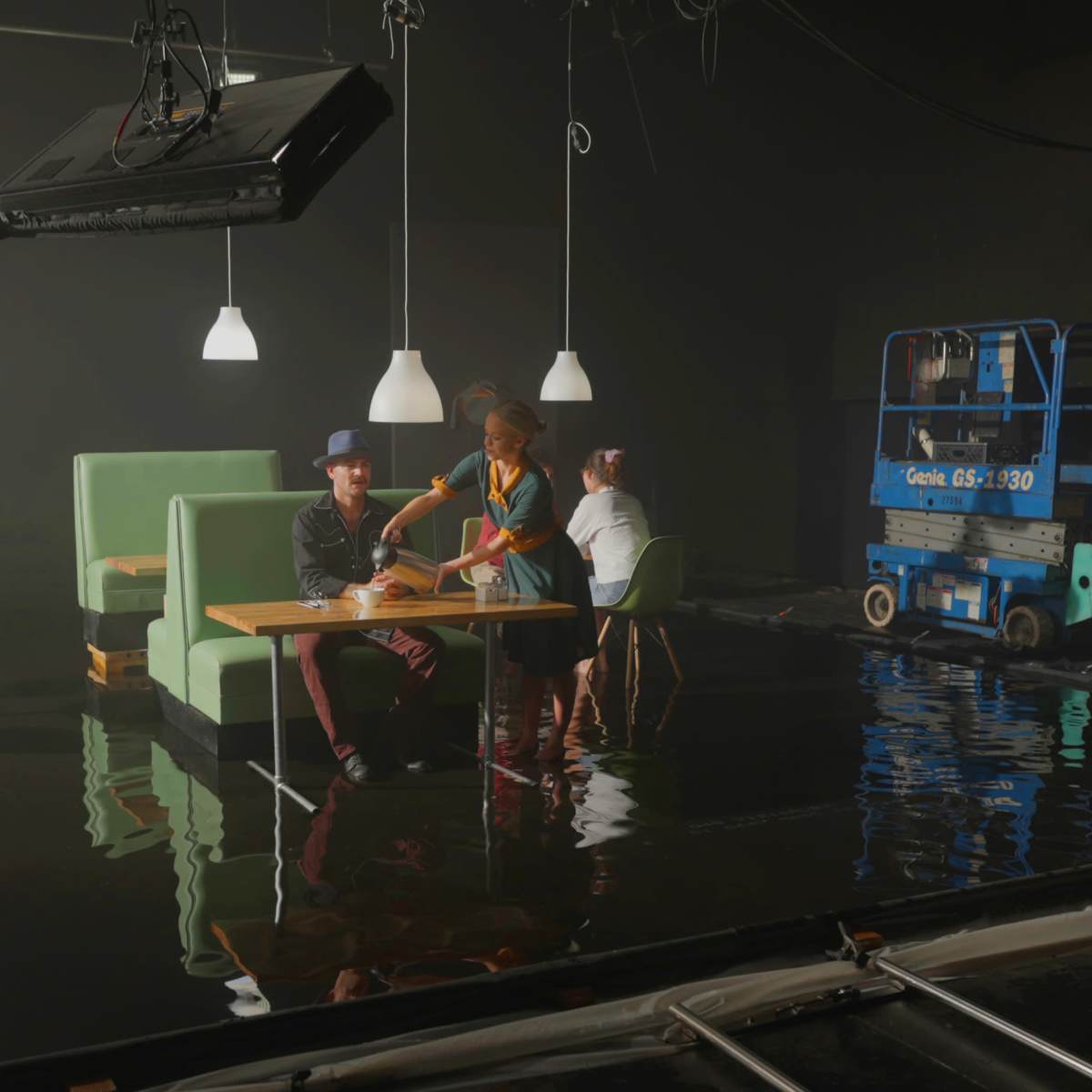 A group of three people sits at a table surrounded by green furniture in a dimly lit studio with water on the floor.