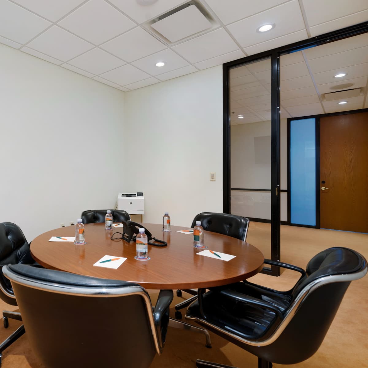 A round wooden conference table is set with four black chairs, water bottles, and notepads in a simple meeting room.