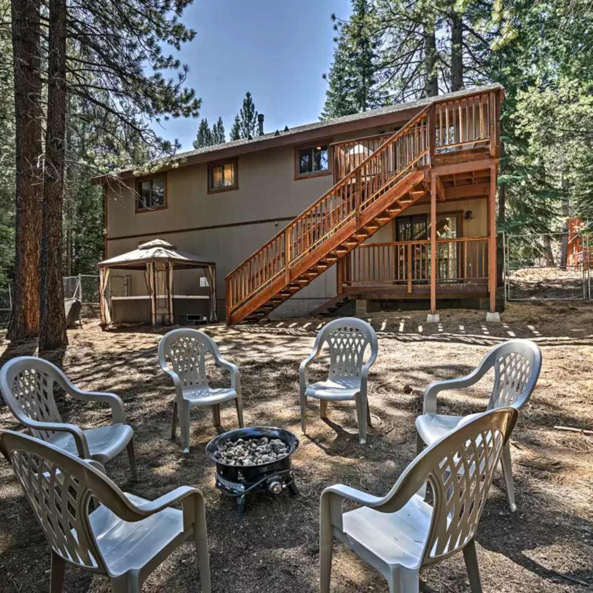 A circular arrangement of white chairs surrounds a fire pit in a forested area, with a two-story house and stairs visible in the background.