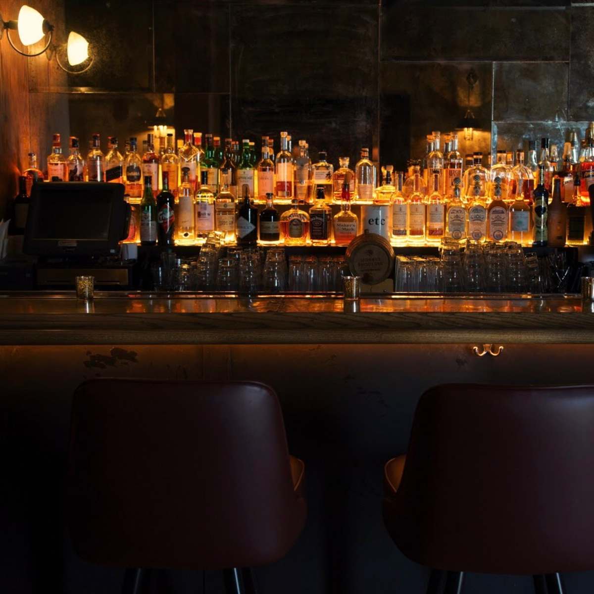 A dimly lit bar with a wide variety of alcoholic bottles displayed on shelves behind a polished counter and two empty bar stools in front.