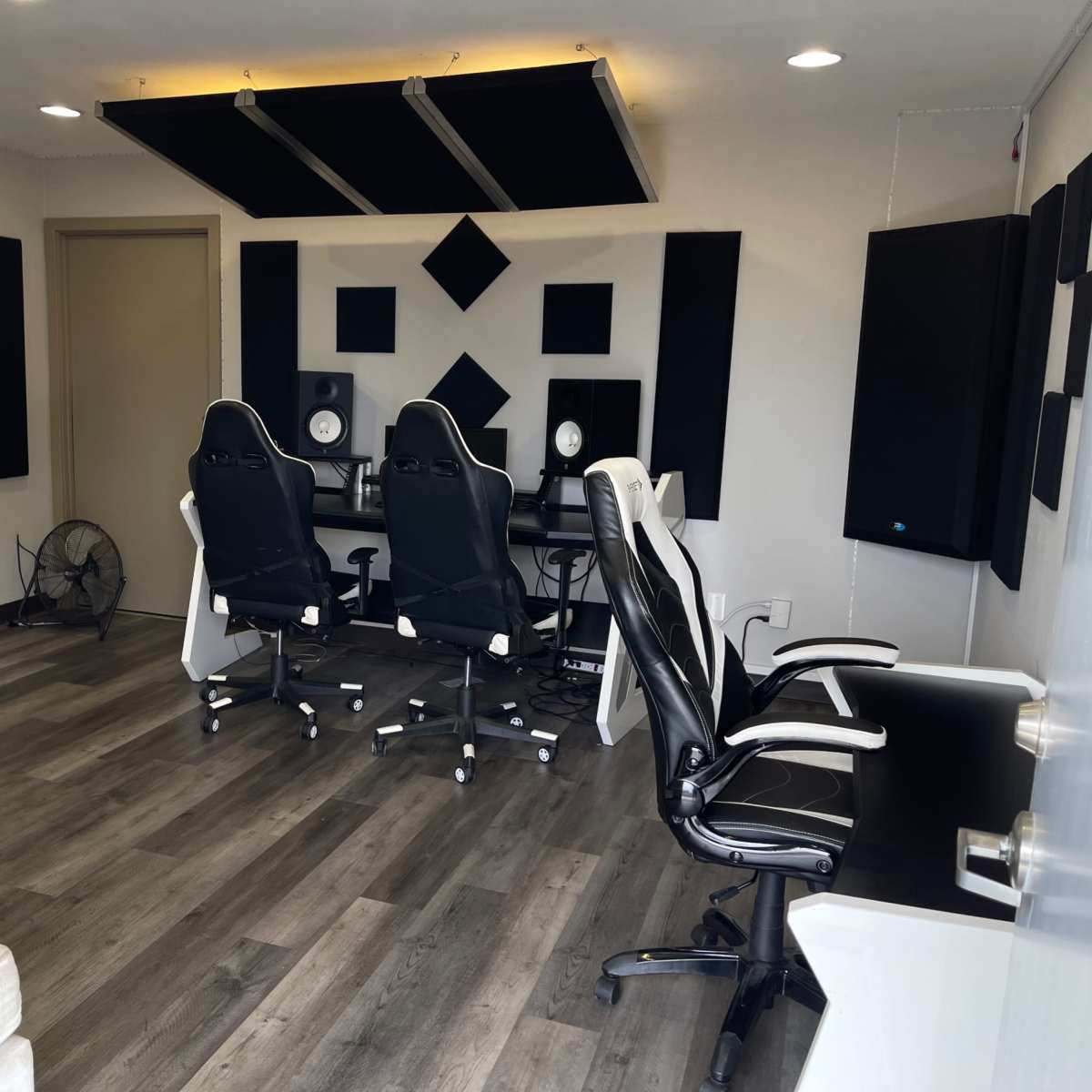 A music studio with two desks, each equipped with audio monitors, surrounded by black acoustic panels on the walls and a fan in the corner.