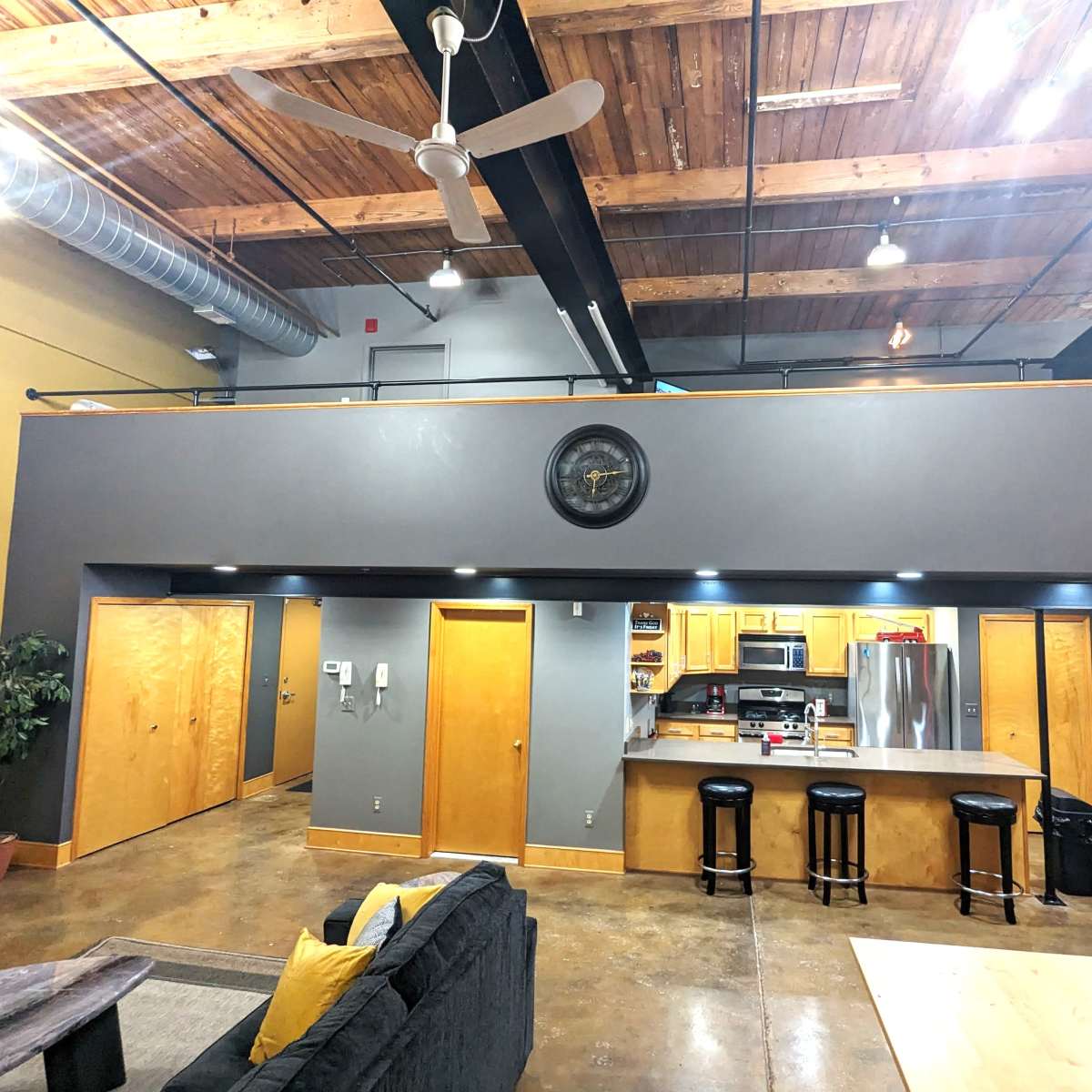 An open-concept loft space with a kitchen, bar stools, and a living area beneath a wooden ceiling.