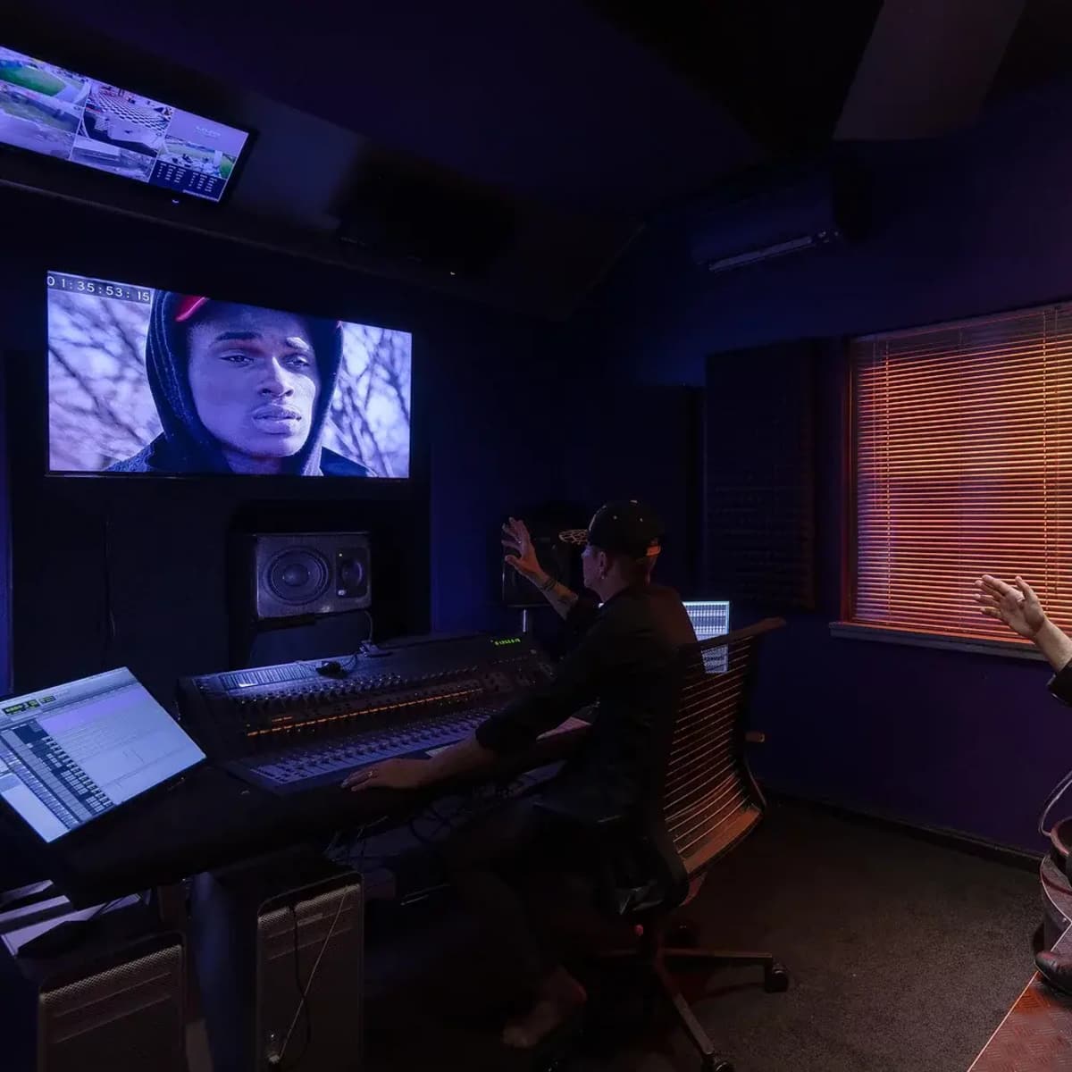 A music production studio features two people, one seated at a mixing console and the other gesturing, while a monitor displays a video of a young man.