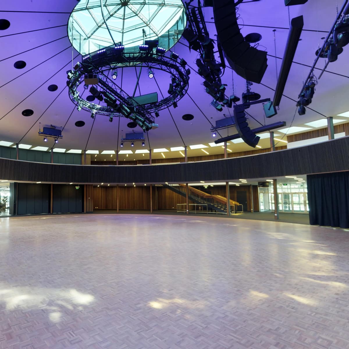 A spacious circular event hall with a glossy wooden floor, large windows, and a modern lighting setup overhead.