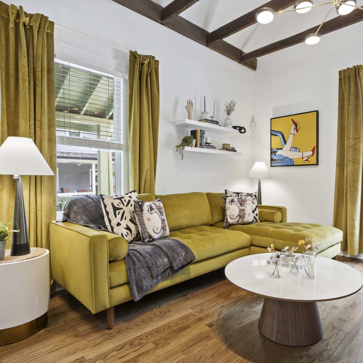 A bright living room features a green sectional sofa with decorative pillows, a round coffee table, and large windows adorned with yellow curtains.
