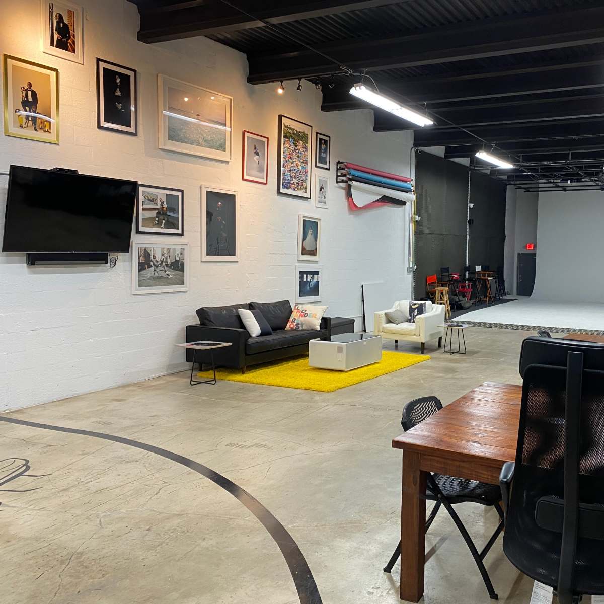 A spacious studio featuring a black sofa, a white armchair, a yellow rug, and a large backdrop area with various studio equipment and framed photographs on the walls.