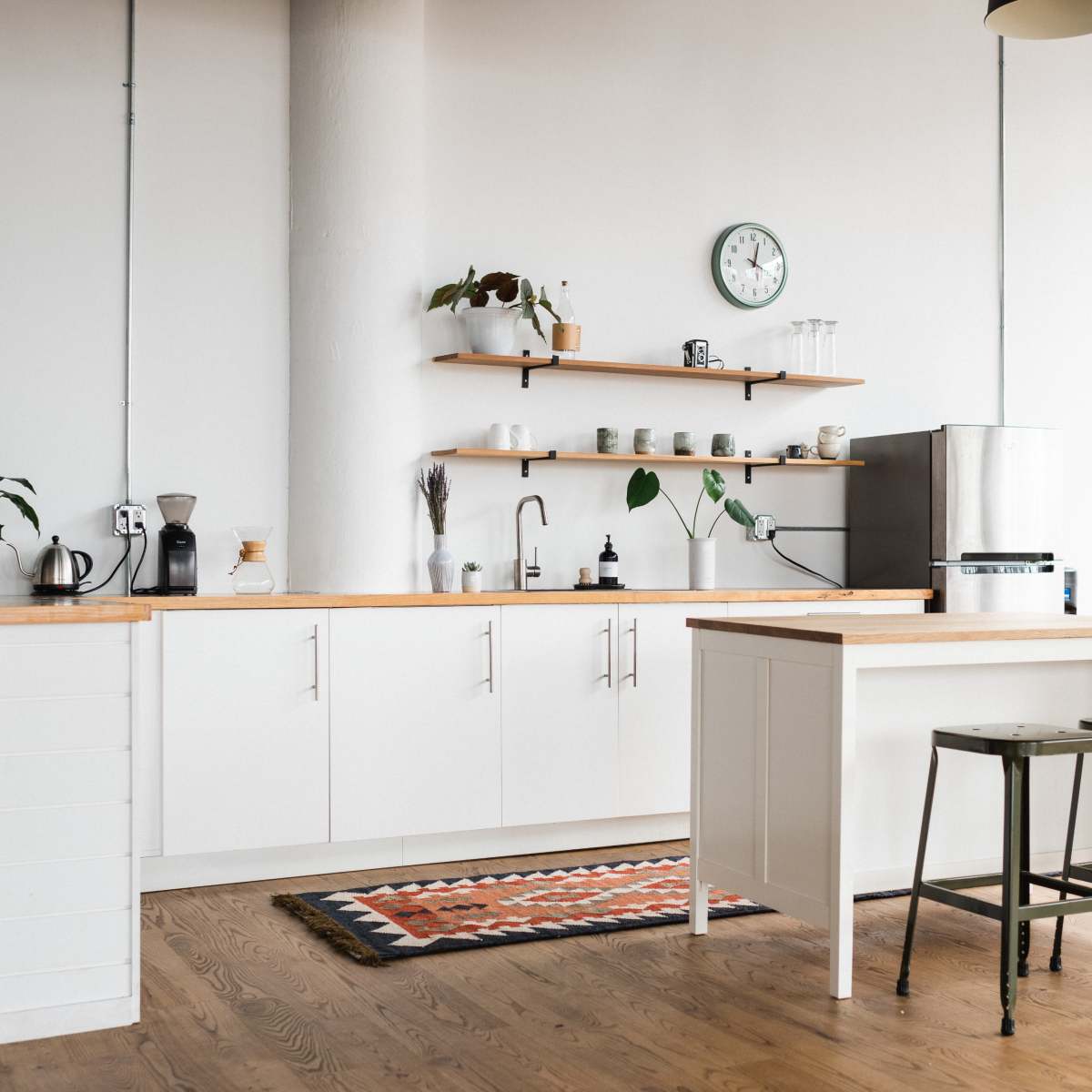 A modern kitchen features white cabinetry, wooden countertops, shelving with plants and dishware, and metal stools on a wooden floor.