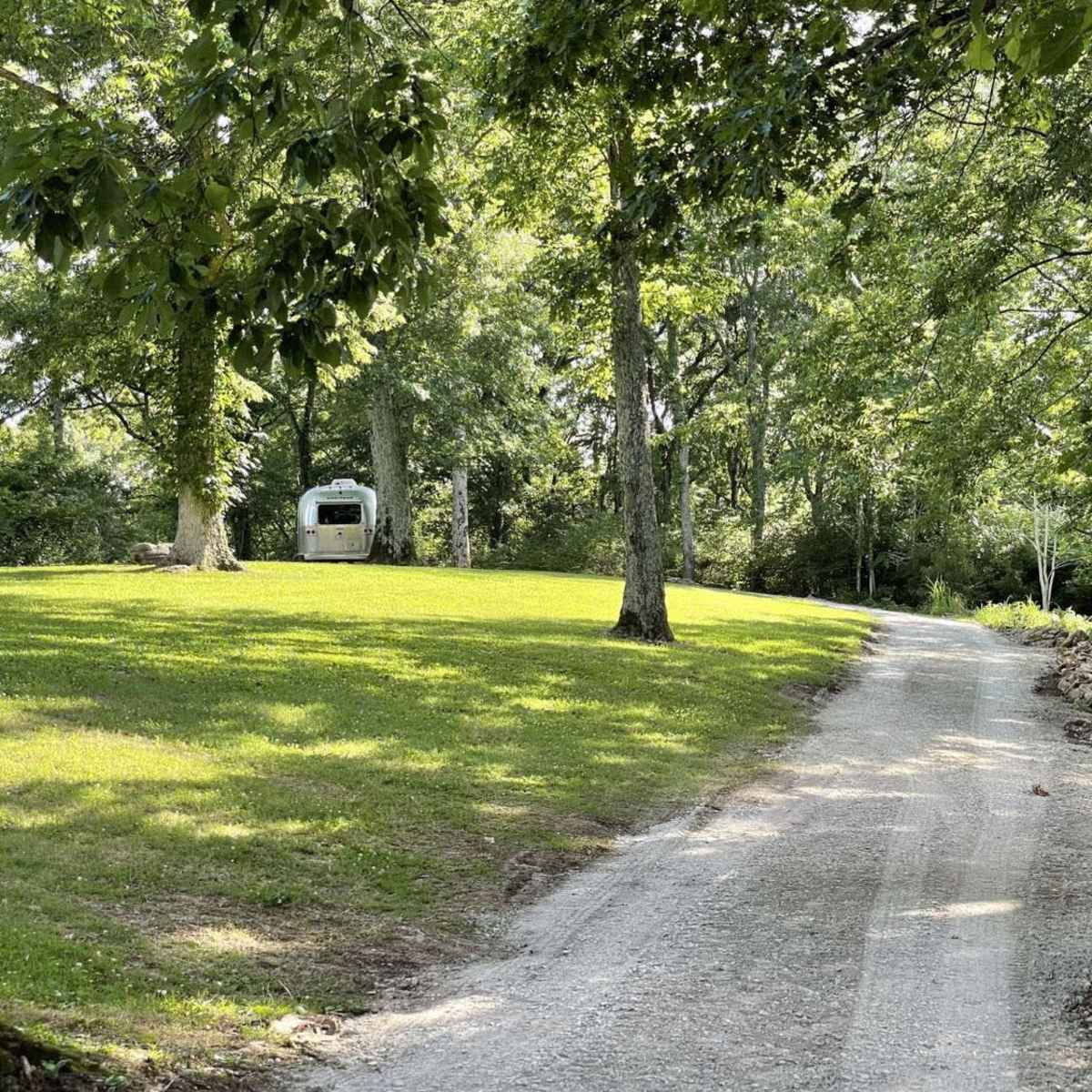 A gravel path winds through a green landscape lined with trees, leading to a small trailer in the distance.