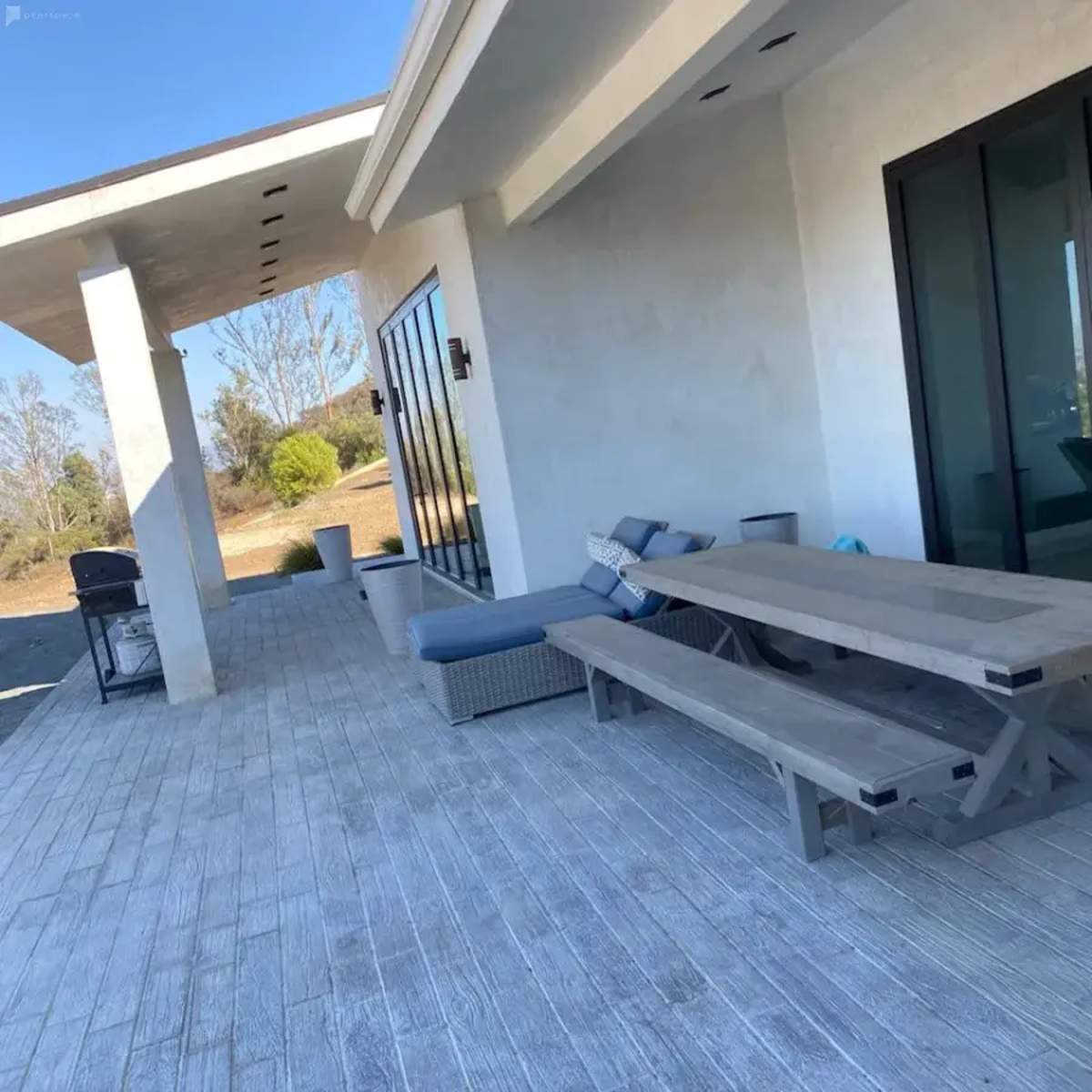 A wooden dining table with benches is placed on a light-colored patio next to large glass doors.