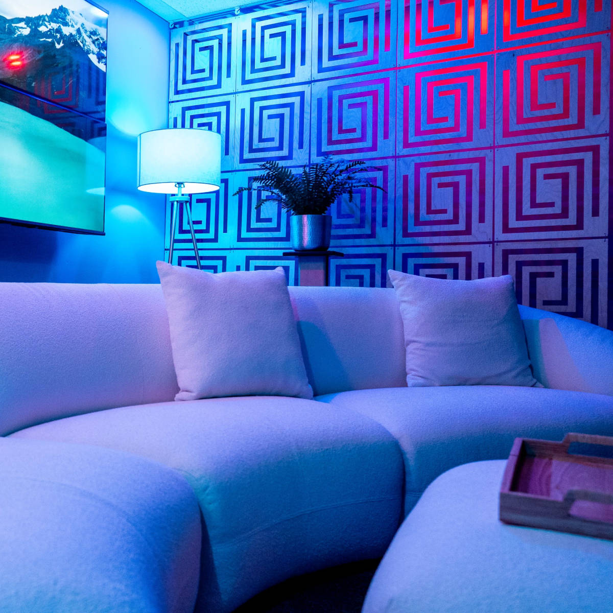 A modern seating area featuring a curved white sofa adorned with decorative pillows, set against a backdrop of geometric wall patterns and illuminated by colored lights.