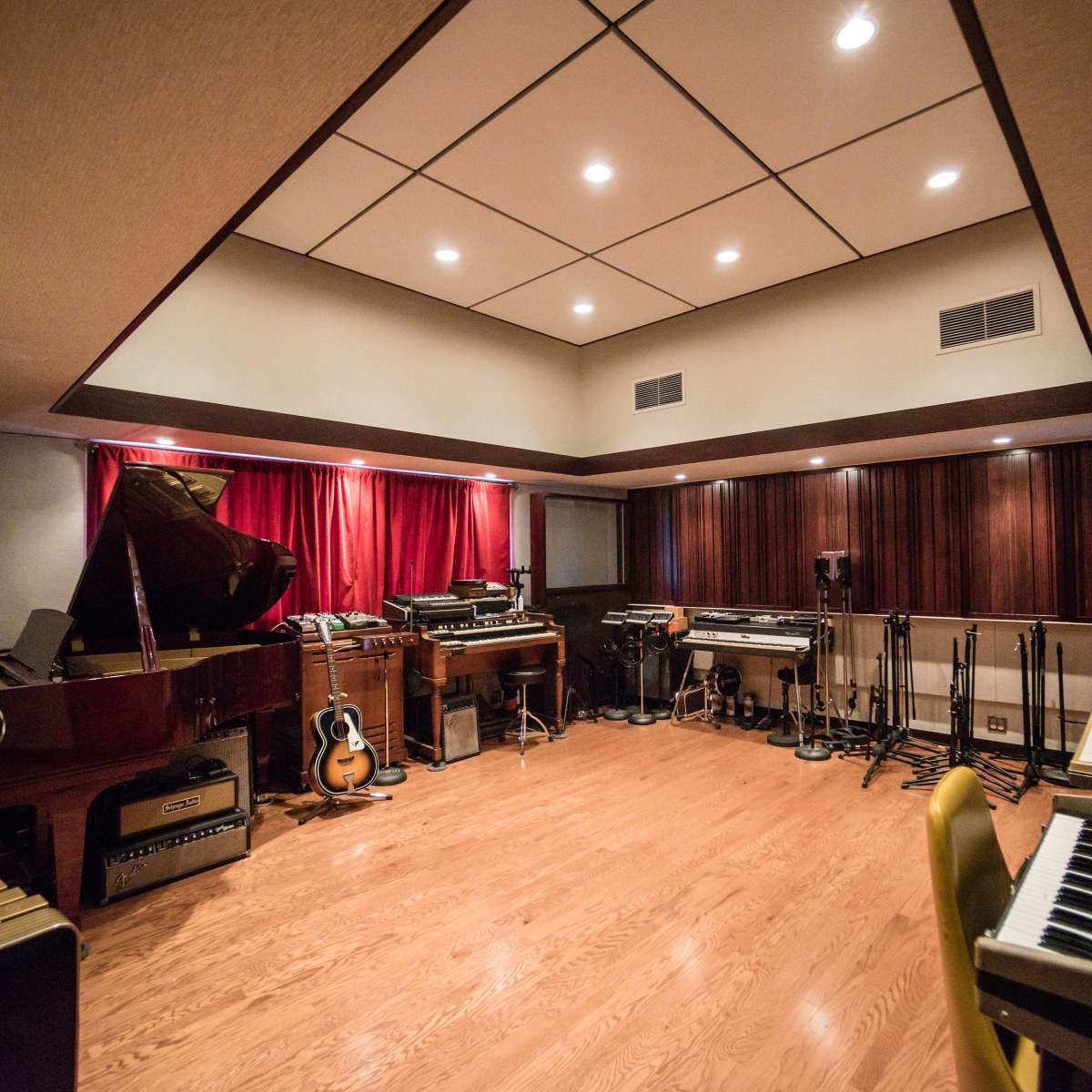 A music studio with various instruments, including a grand piano, guitars, and synthesizers, arranged within a room featuring wooden walls and red drapes.