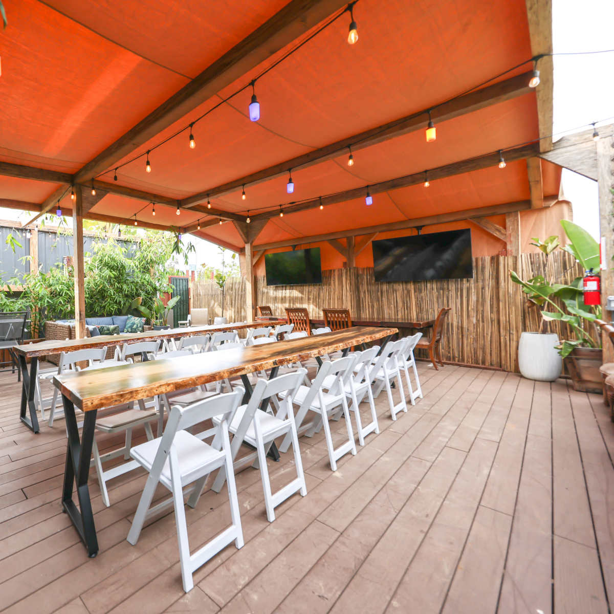 An outdoor dining area with a long wooden table surrounded by white chairs, covered by an orange canopy and illuminated by string lights.