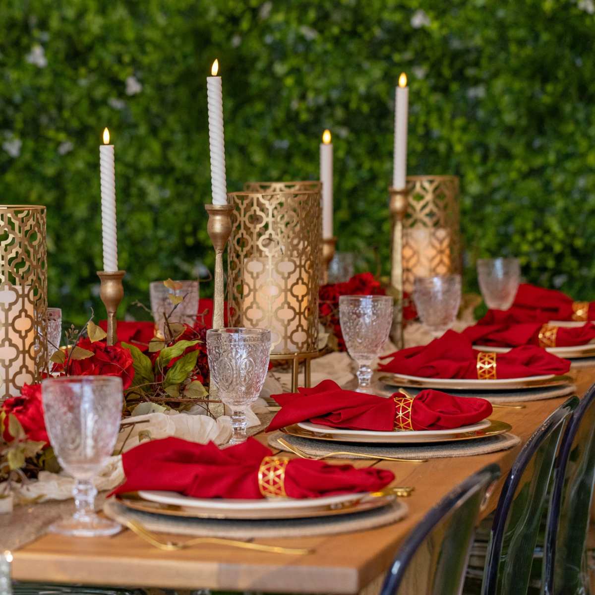 A beautifully arranged dining table features elegant golden candle holders, red napkins, crystal glasses, and a backdrop of lush greenery.