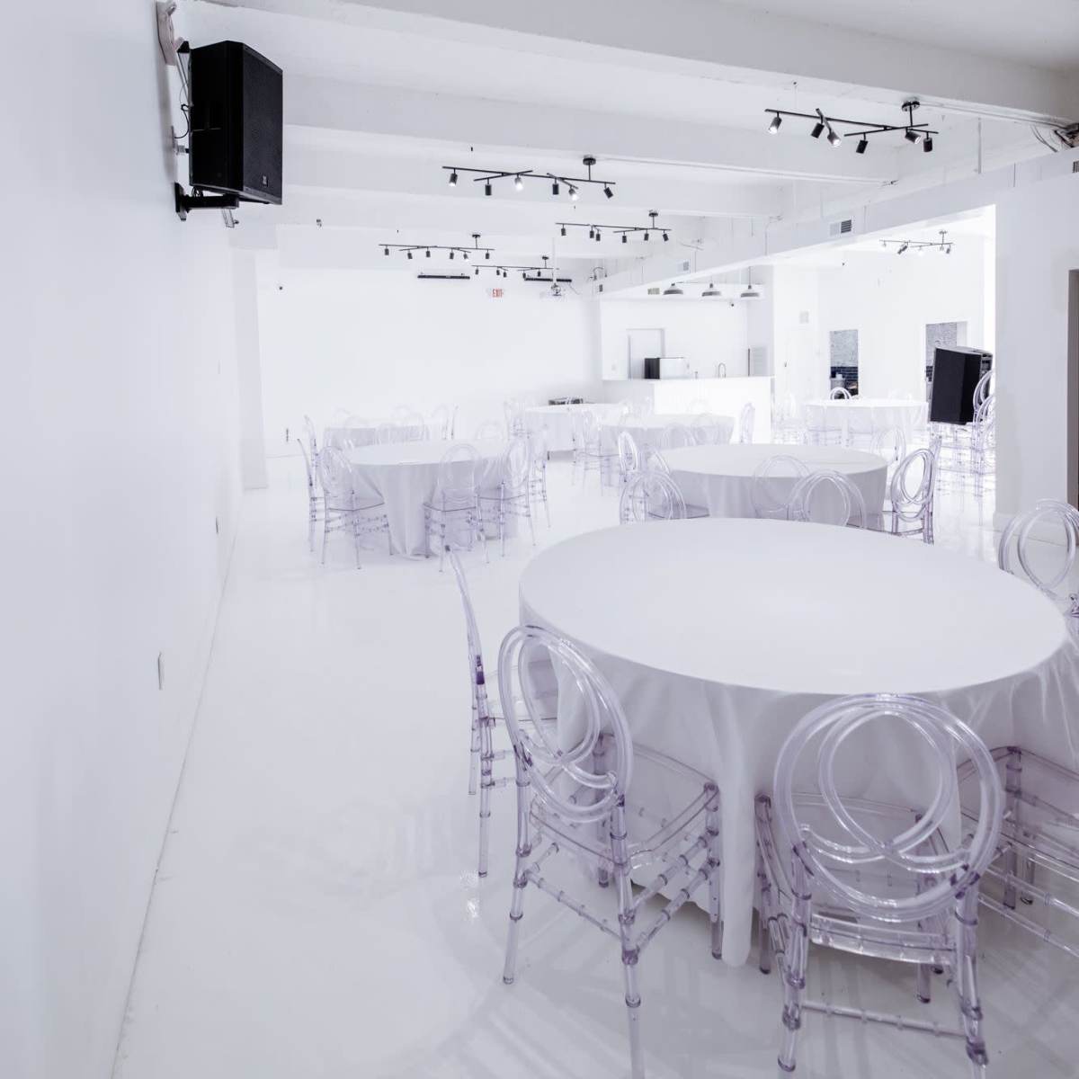 A spacious event venue features several round tables with transparent chairs set against a white backdrop.