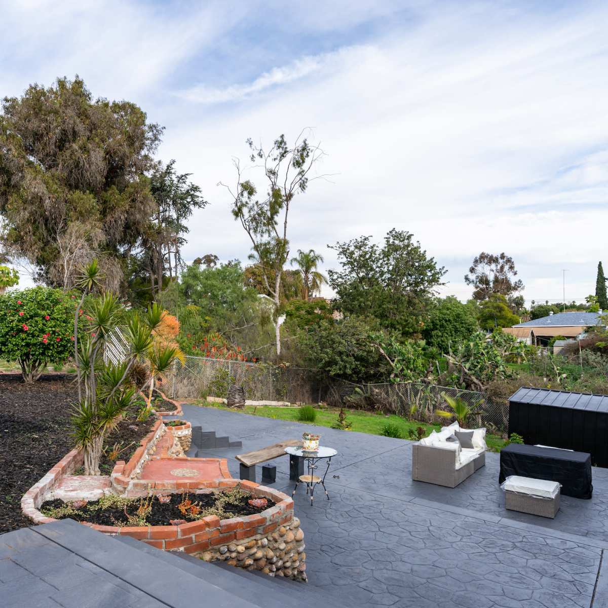 A backyard features a patio with gray furniture, a stone pathway, and landscaped greenery in the background.