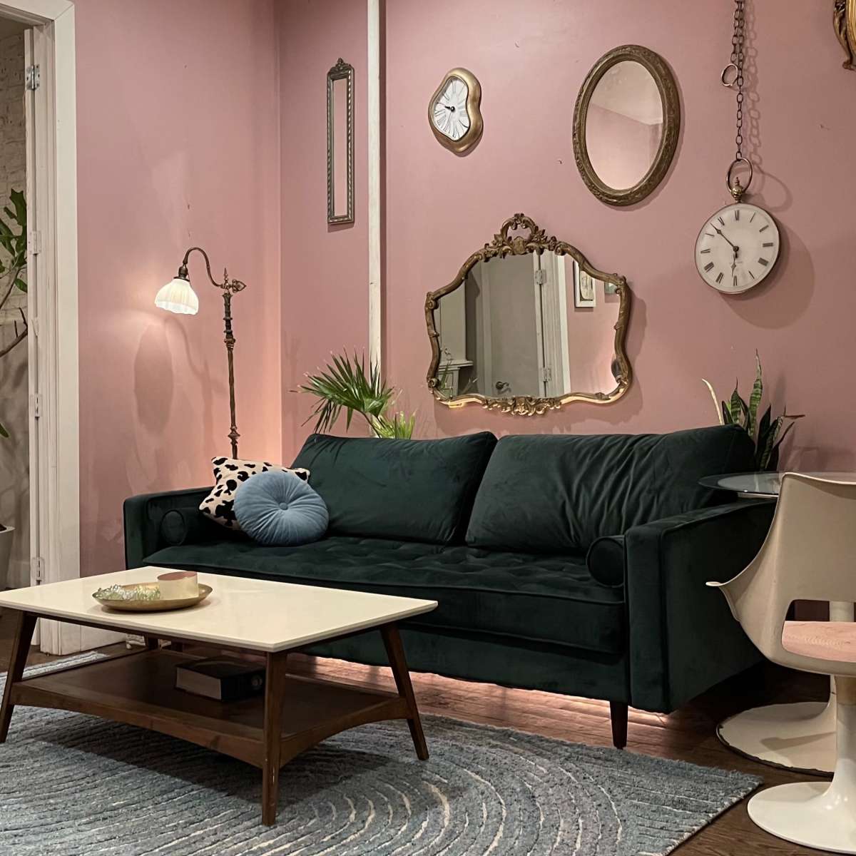The scene features a cozy living room with a green velvet sofa, a light wooden coffee table, and stylish chairs surrounding a small dining table, all against a backdrop of pink walls adorned with various mirrors.