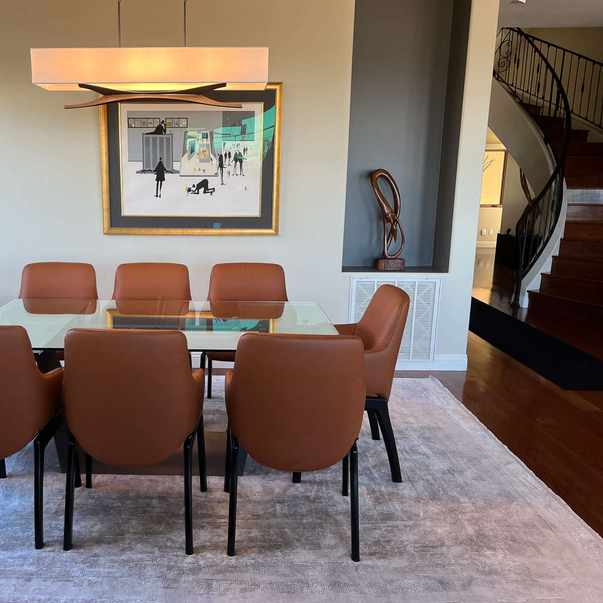 A spacious dining room features a glass table surrounded by eight brown leather chairs, with an artistic painting on the wall and a modern light fixture above.