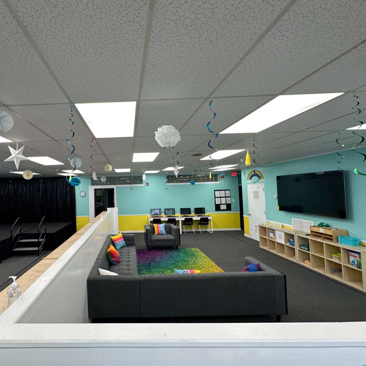A bright, spacious room with a seating area featuring a gray couch, a small stage at one end, and colorful decorations hanging from the ceiling.