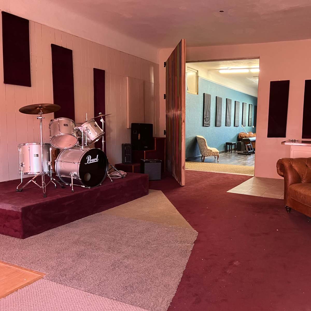 A music practice room featuring a drum set on a raised platform, a brown leather couch, and a doorway leading to a blue-walled space with chairs and recording equipment.