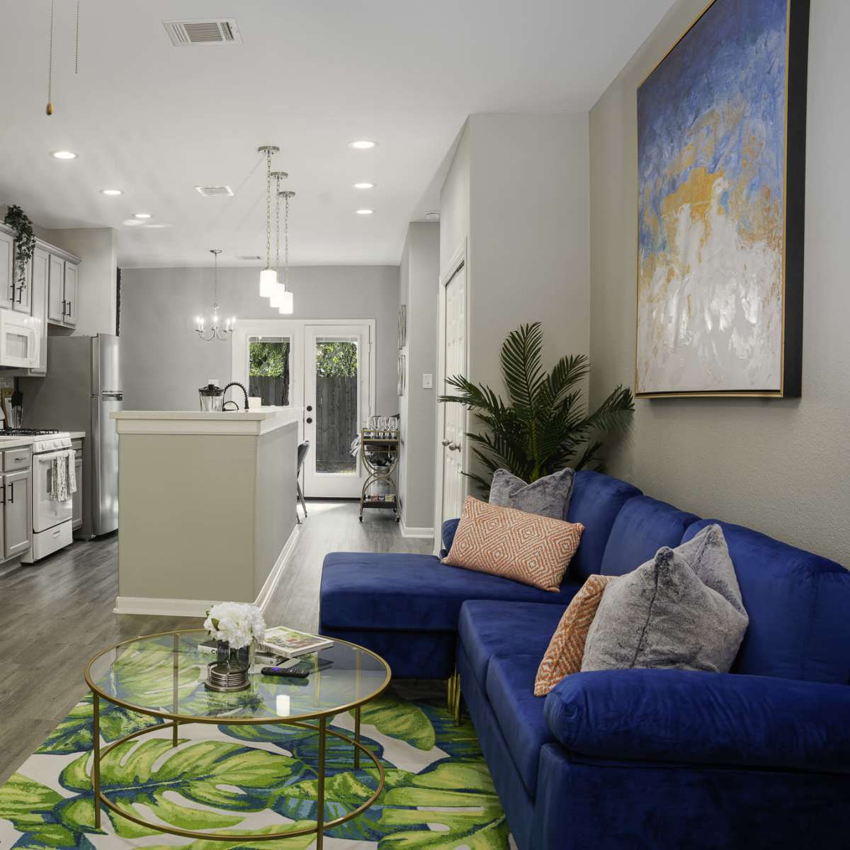 A modern living room features a blue couch, a glass coffee table, and a small workspace, with an open kitchen visible in the background.