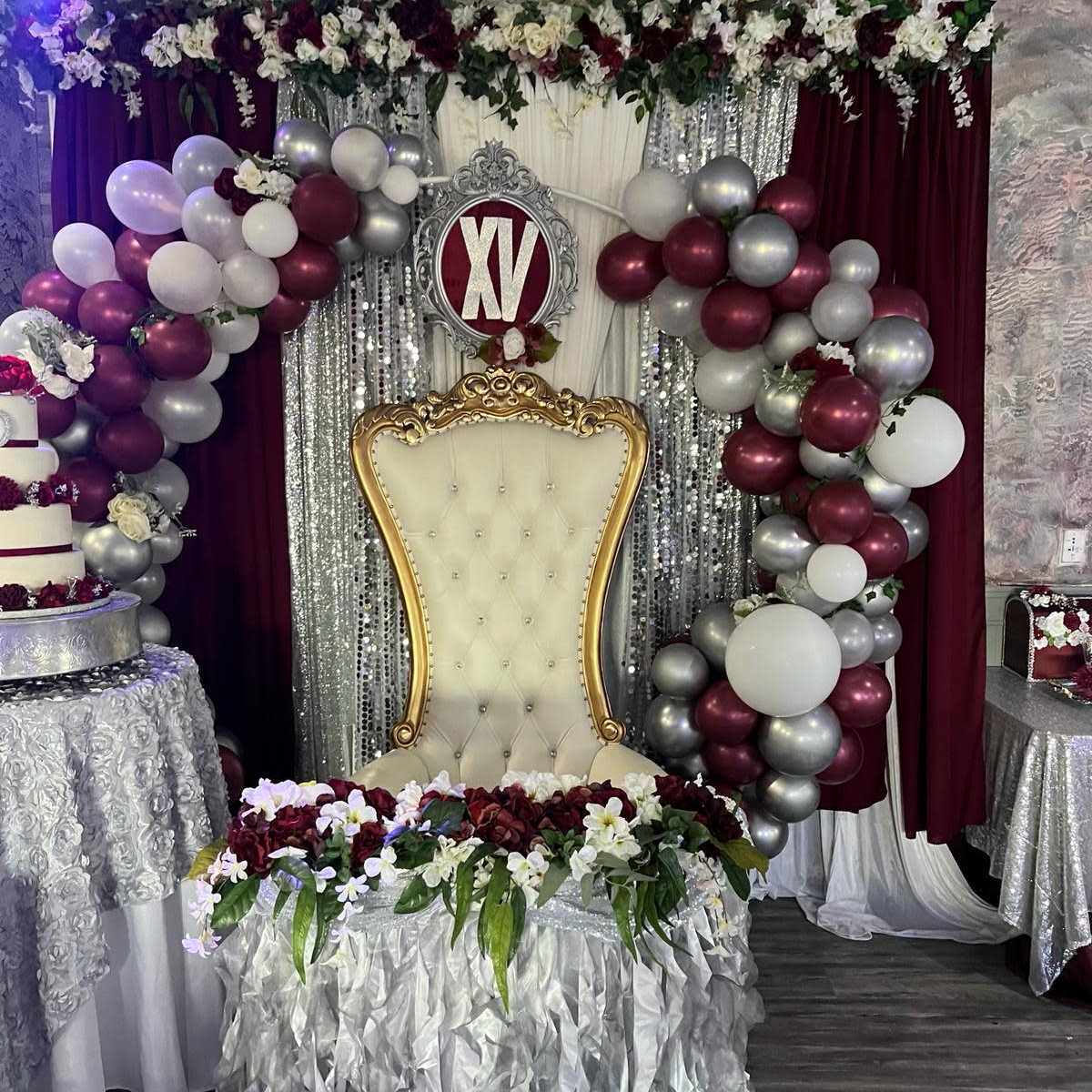 A decorated event space with a lavish throne chair surrounded by a floral and balloon arch, complete with a "XV" sign above it.