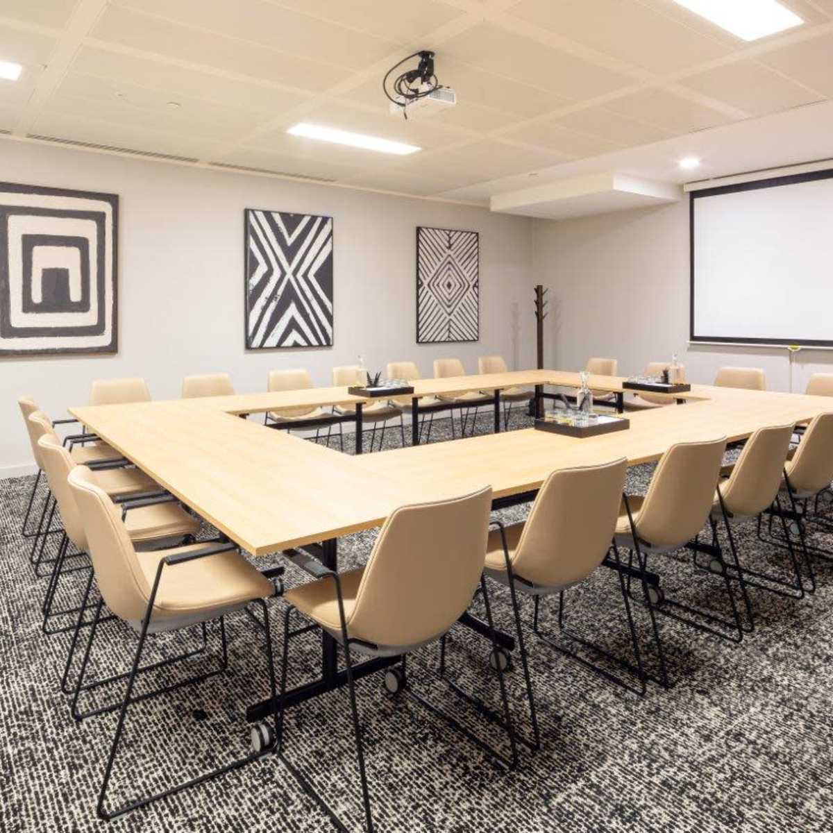A rectangular conference room is set up with a large wooden table surrounded by beige chairs, featuring artwork on the walls and a projector screen at one end.