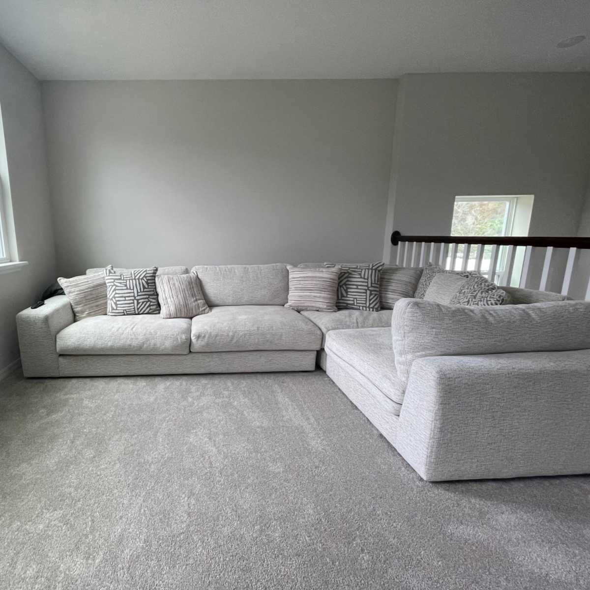A spacious living area features a light-colored sectional sofa against a grey carpet with large windows allowing natural light.