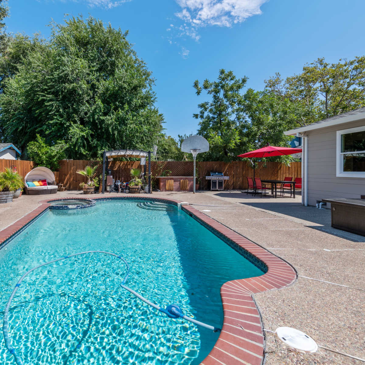 A swimming pool is surrounded by a patio area with furniture and greenery in a backyard setting.