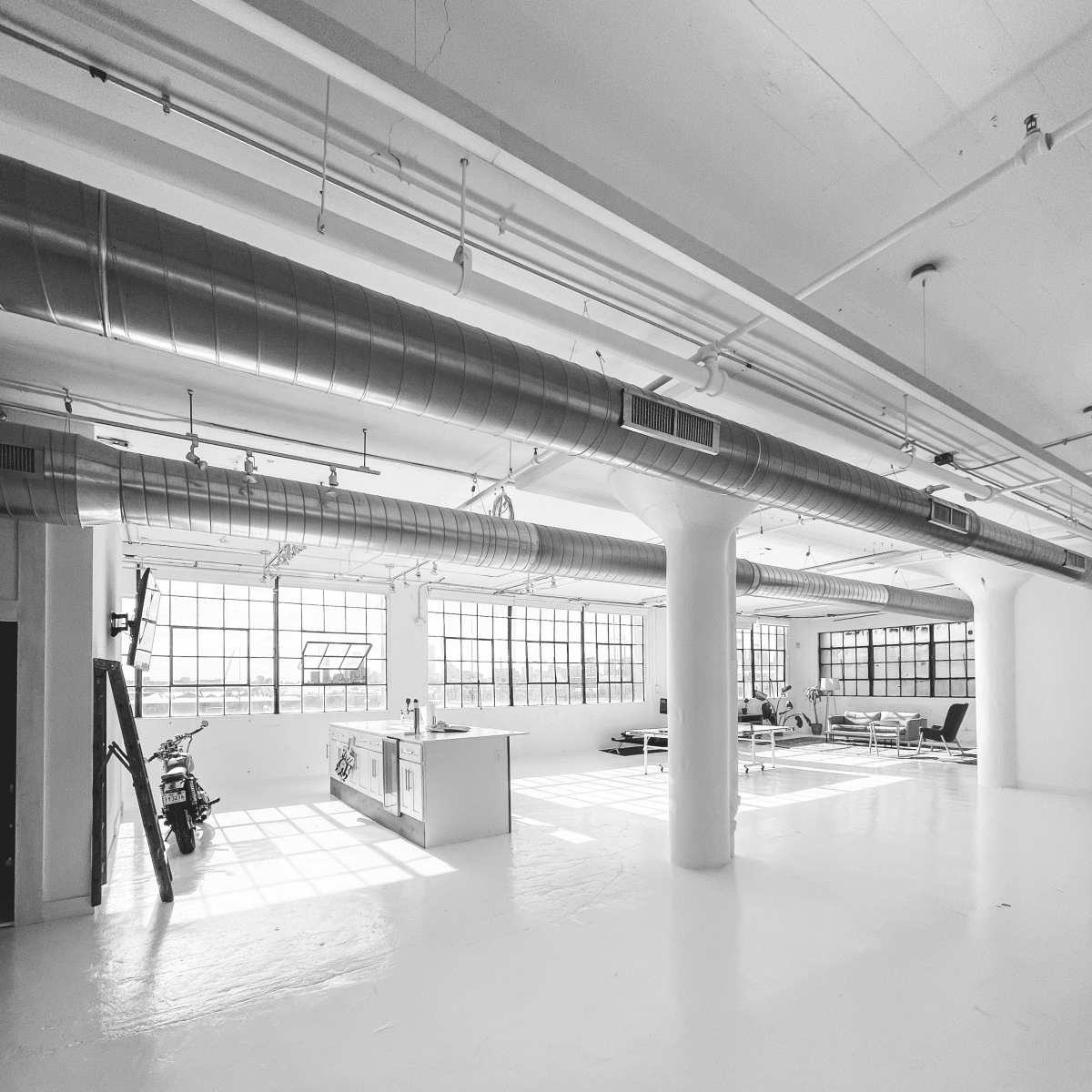 A spacious, industrial-style room with large windows, exposed ductwork, and minimal furnishings.