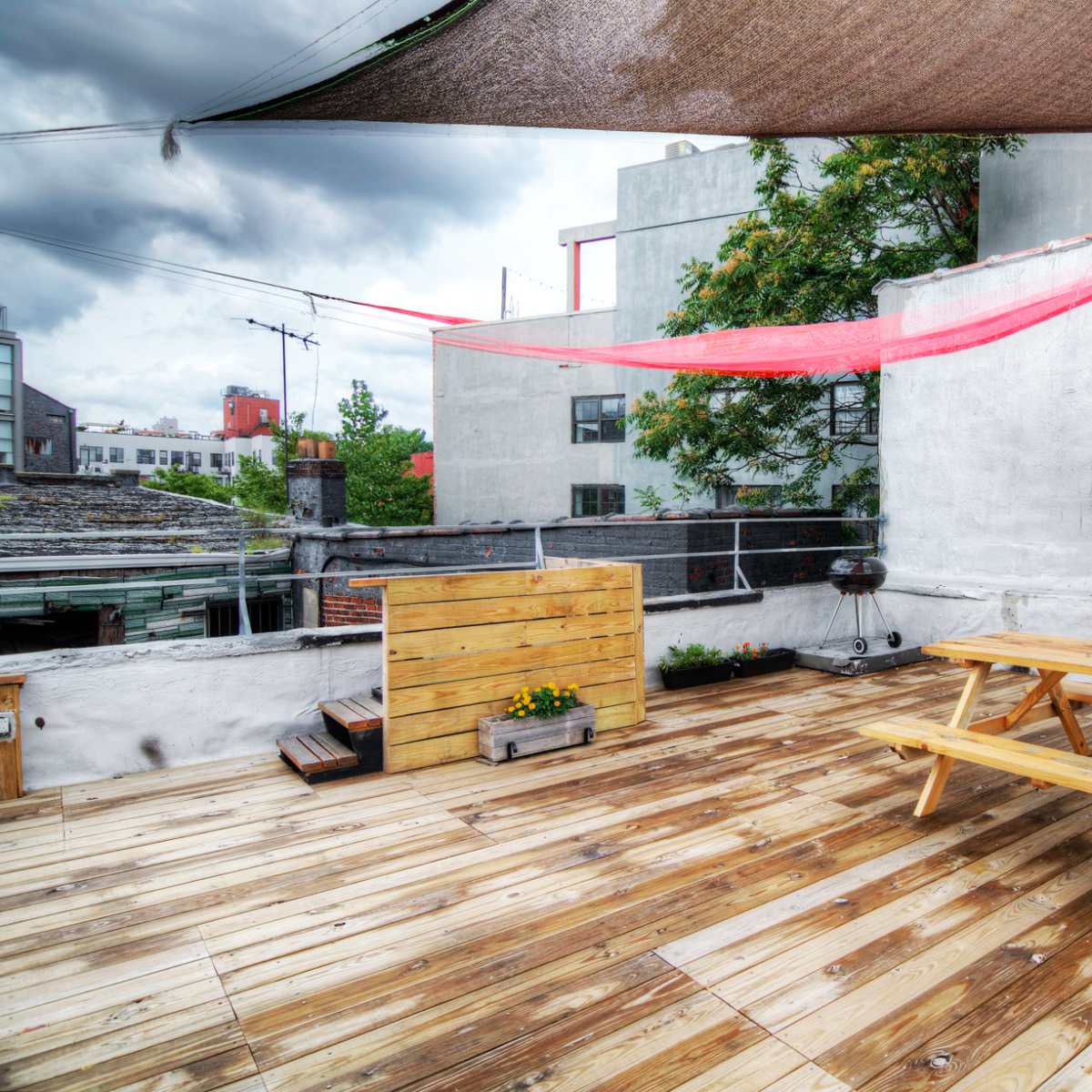 A rooftop deck with wooden flooring, a picnic table, and shaded areas created by large fabric drapes, surrounded by buildings and greenery.