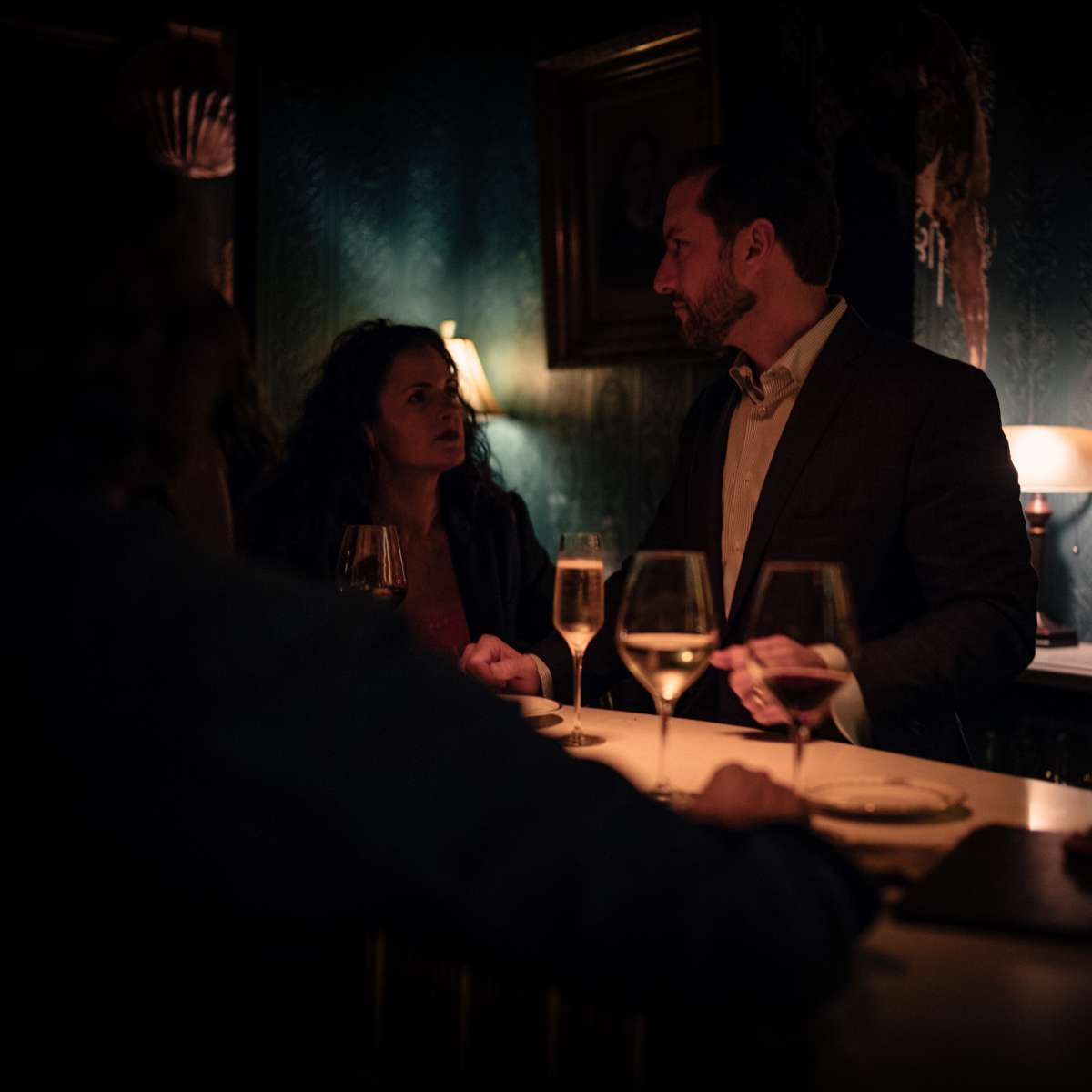A man and a woman are engaged in conversation at a dimly lit bar, with glasses of champagne and wine on the countertop.