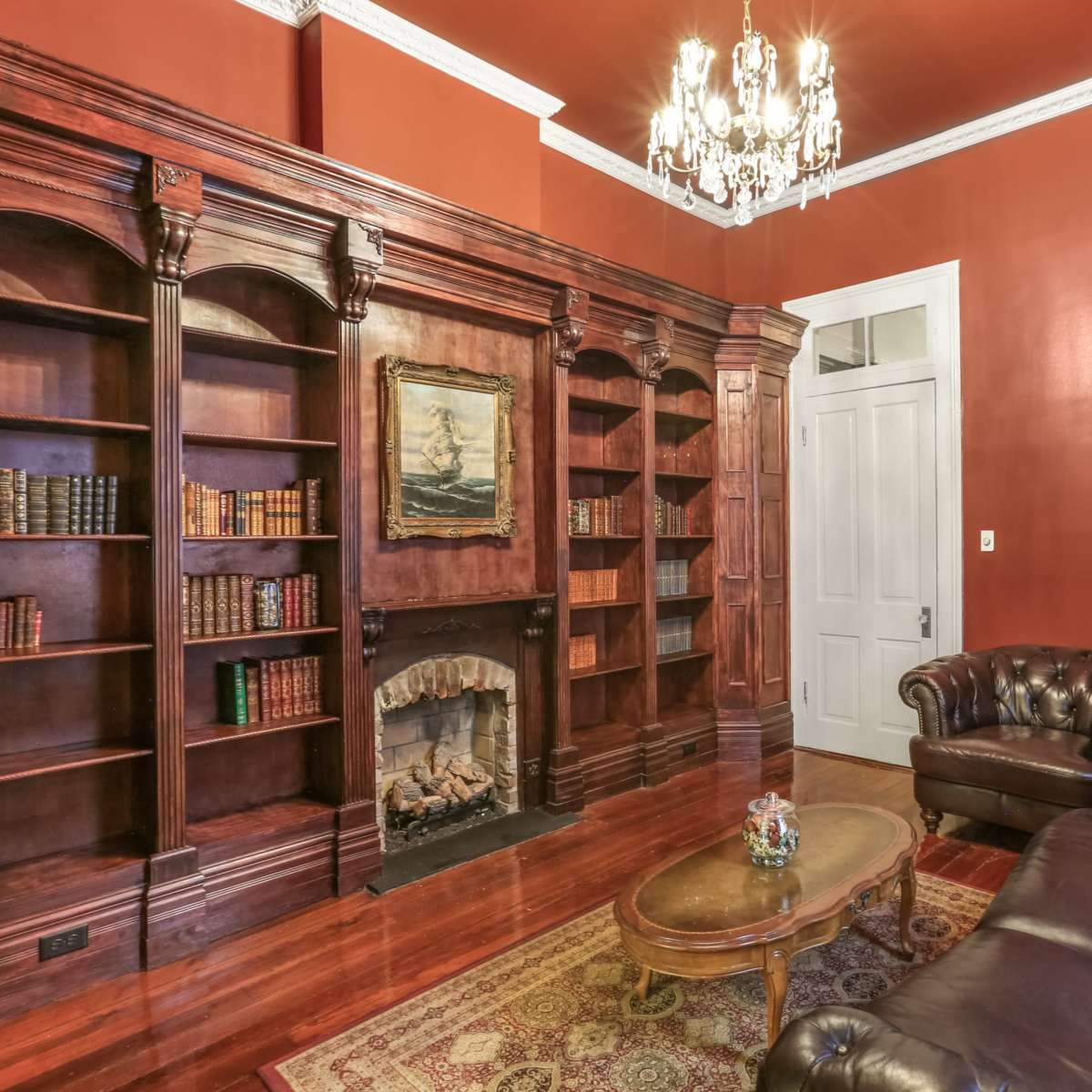 A richly furnished room with dark wood bookshelves, a leather chesterfield sofa, and a decorative chandelier against a red wall.