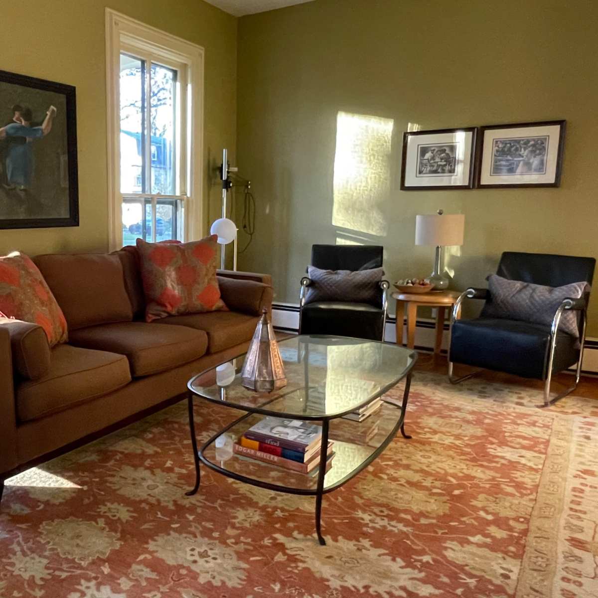 A cozy living room features a brown sofa with decorative pillows, a glass coffee table, and two armchairs, all set against green walls and illuminated by natural light.