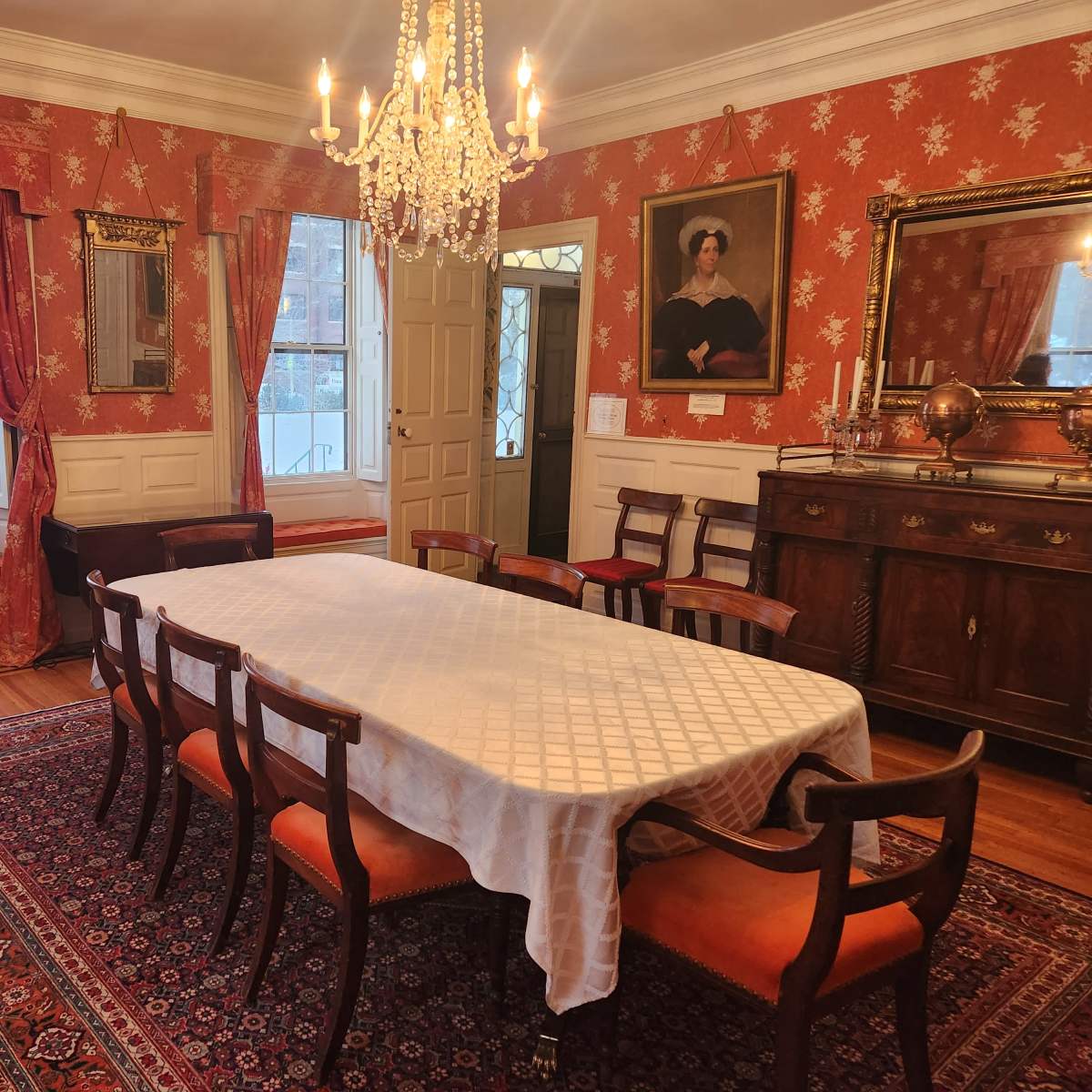 The dining room features a long, white table set with a tablecloth, surrounded by eight wooden chairs, with patterned wallpaper and a chandelier hanging from the ceiling.